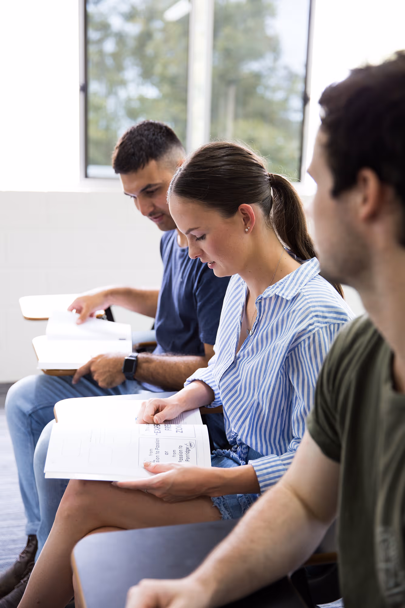 Three young adults sitting in a classroom, reading and studying from open books.