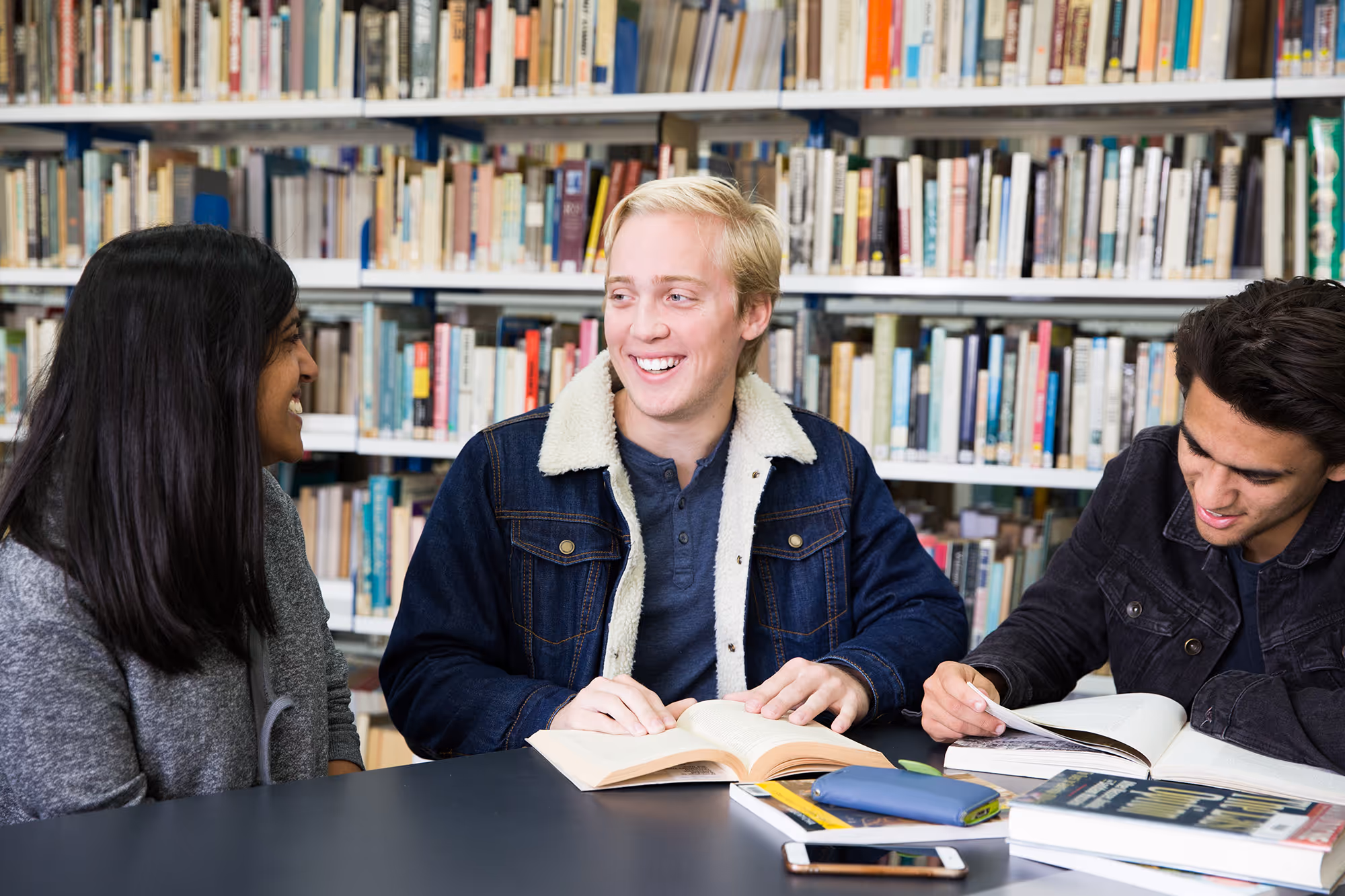 Three college students studying and talking at a library table with books and a smartphone.