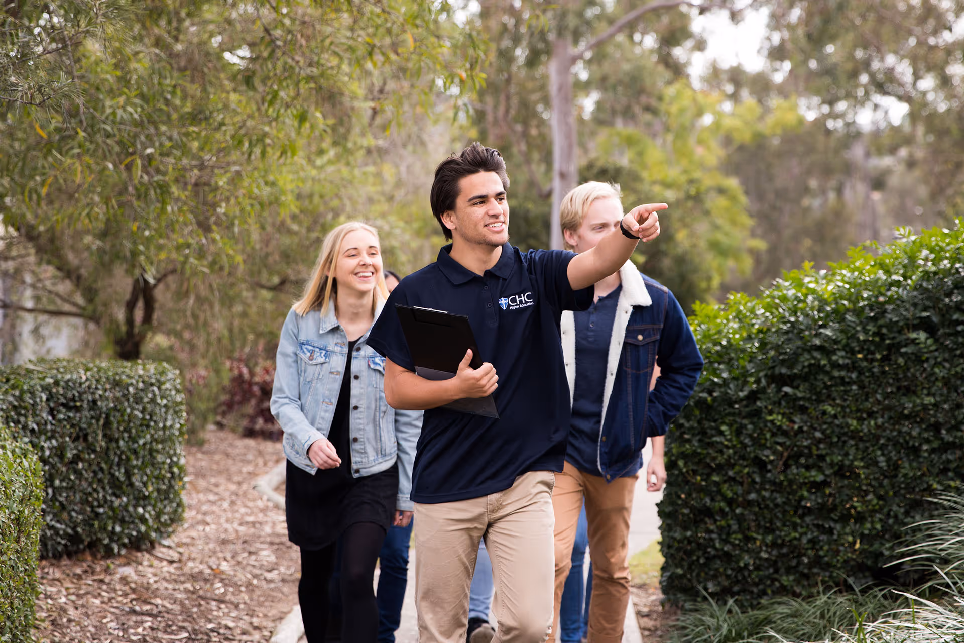 A young man in a CHC polo shirt holding a clipboard points ahead while walking outdoors with smiling friends around him.