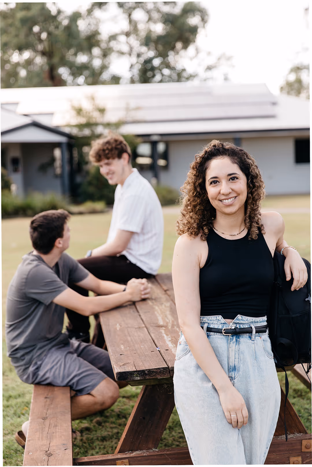 Smiling young woman in a black sleeveless top and light jeans leaning on a wooden picnic table with two young men sitting and talking in the background outdoors.