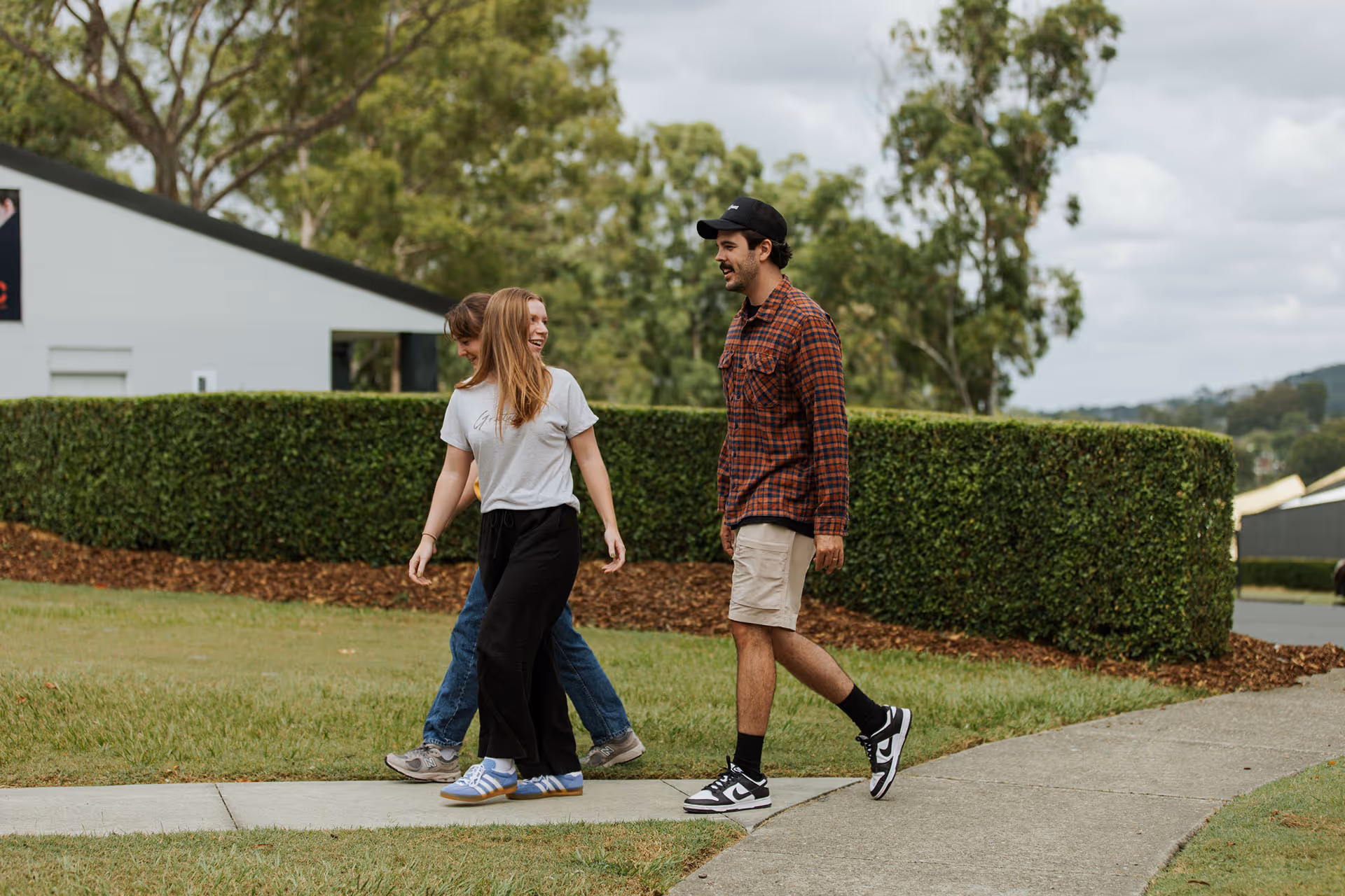 Three young adults walking and chatting on a sidewalk near trimmed hedges and green trees on a cloudy day.