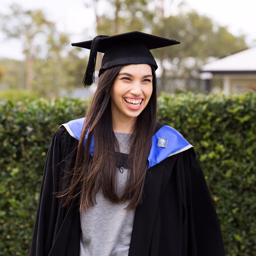 Smiling graduate wearing a black cap and gown with a blue academic stole outdoors.