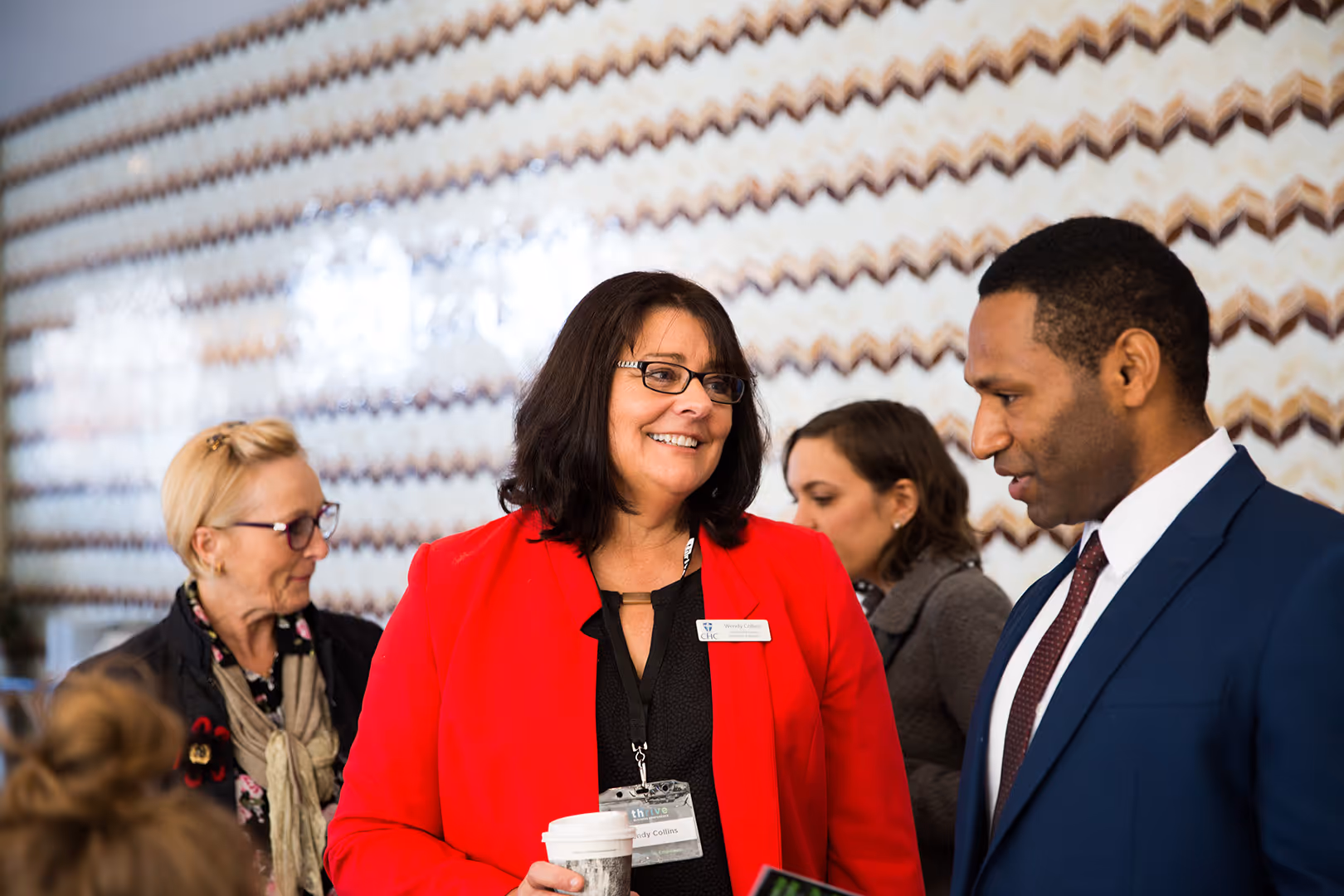 Two professionals, a woman in a red blazer holding a coffee cup and a man in a blue suit, engaged in conversation at a business event.