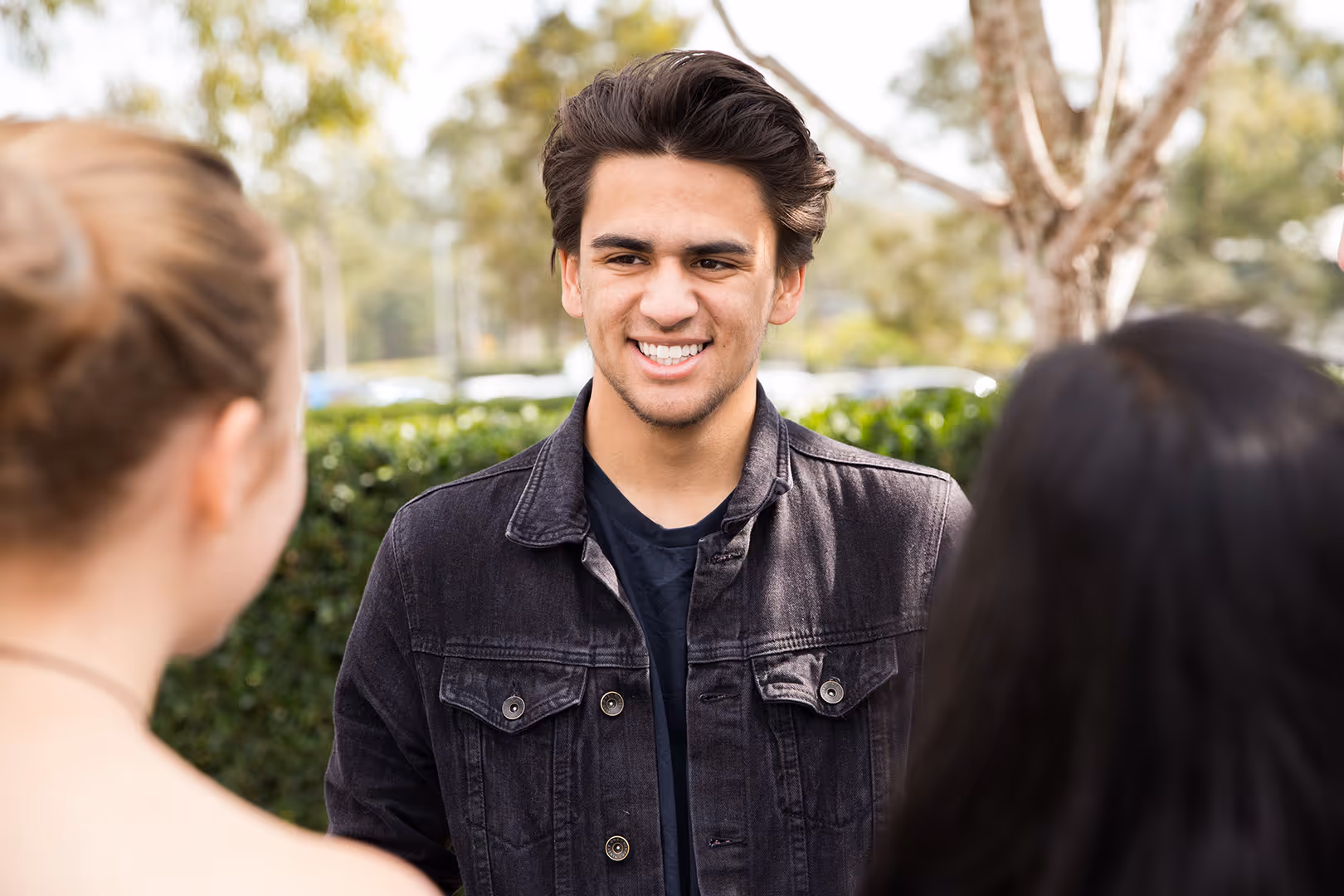 Smiling young man wearing a black denim jacket talking to two people outdoors.