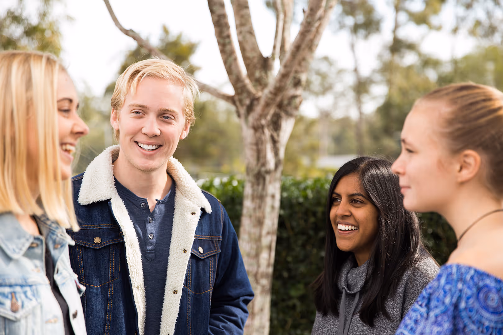 Four young adults standing outside, smiling and engaging in conversation with trees and greenery in the background.