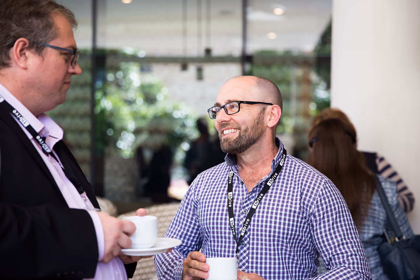 Two men wearing glasses and lanyards holding white coffee cups and engaging in conversation at a social event.