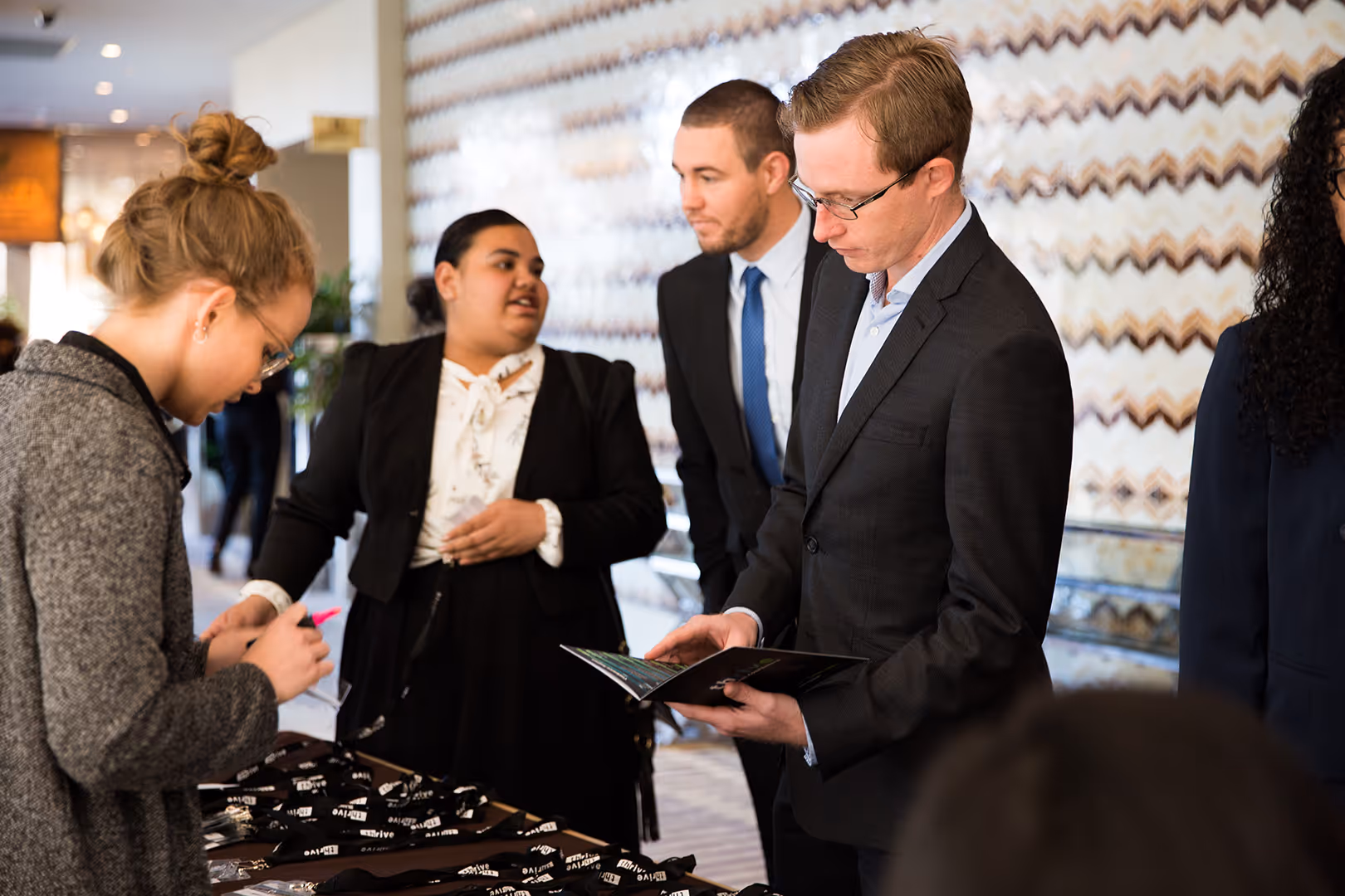 Four business professionals interacting at an event, one reading a booklet, another writing with a pink marker, and two conversing.