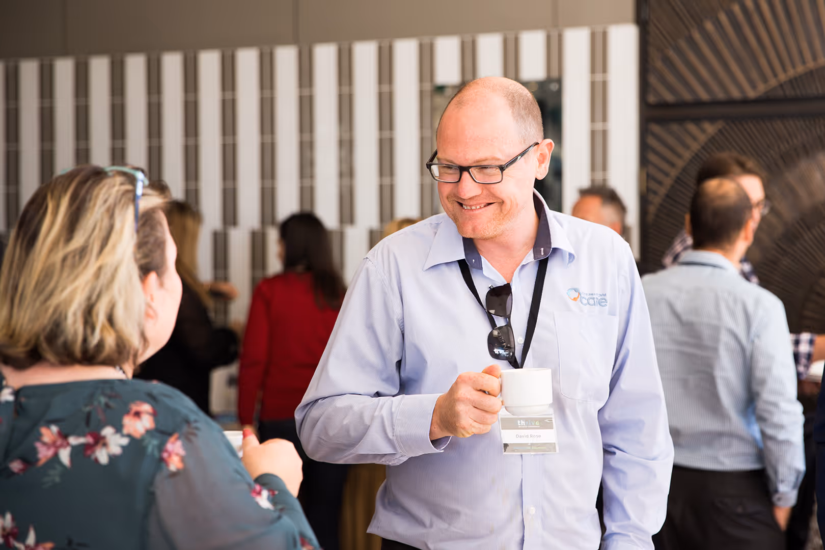 Man in glasses and light blue shirt holding a coffee cup and smiling while talking to a woman at a social gathering.