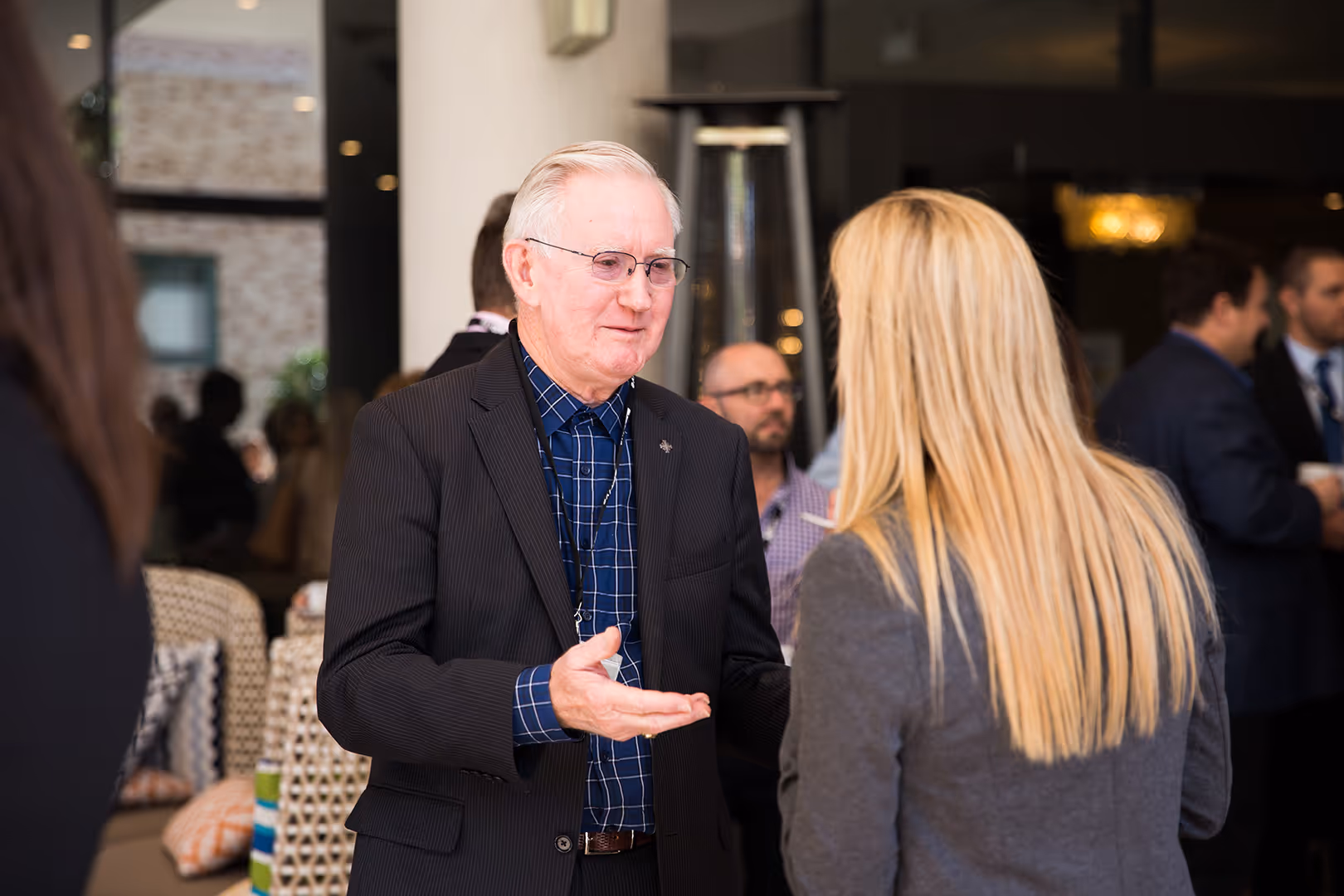 Older man in glasses and a dark suit talking to a blonde woman with long hair in a business setting.