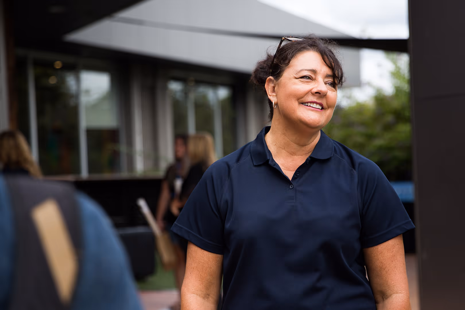 Smiling woman in a navy blue polo shirt standing outdoors with blurred people and building in the background.