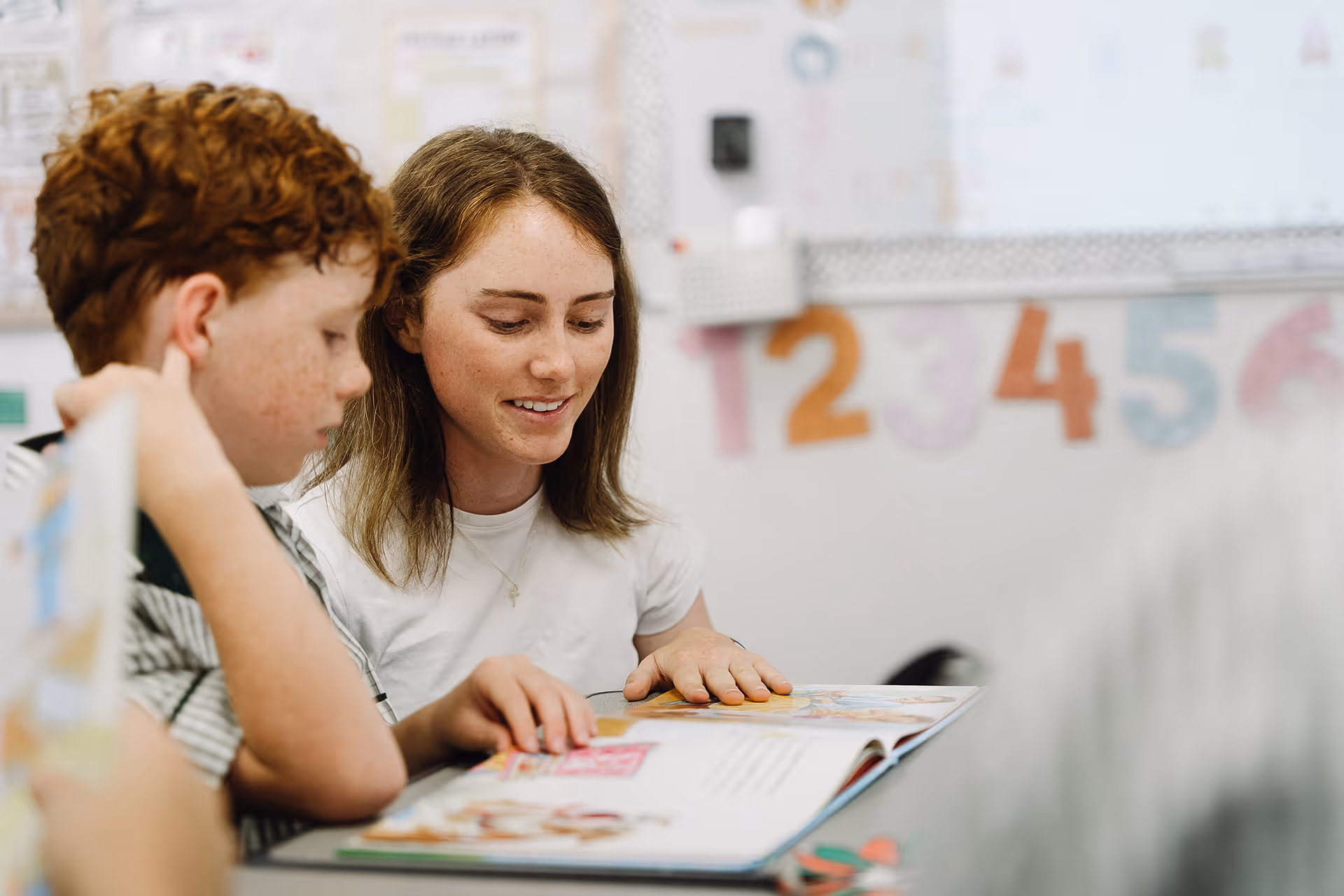 Teacher reading a book with a young student in a classroom decorated with colorful numbers.