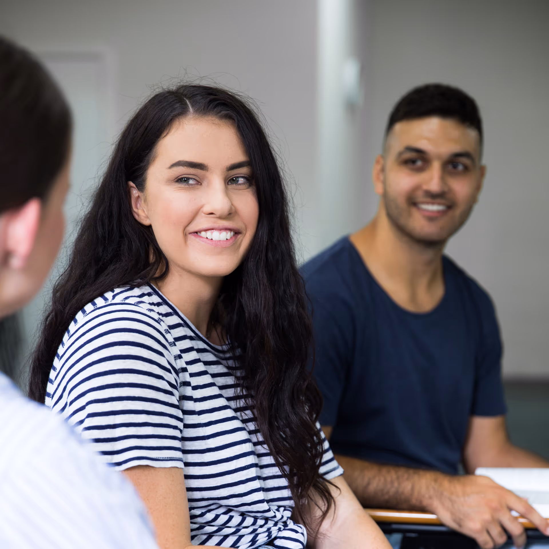 Smiling young woman in a striped shirt looking at a person while a man in a navy shirt smiles in the background.