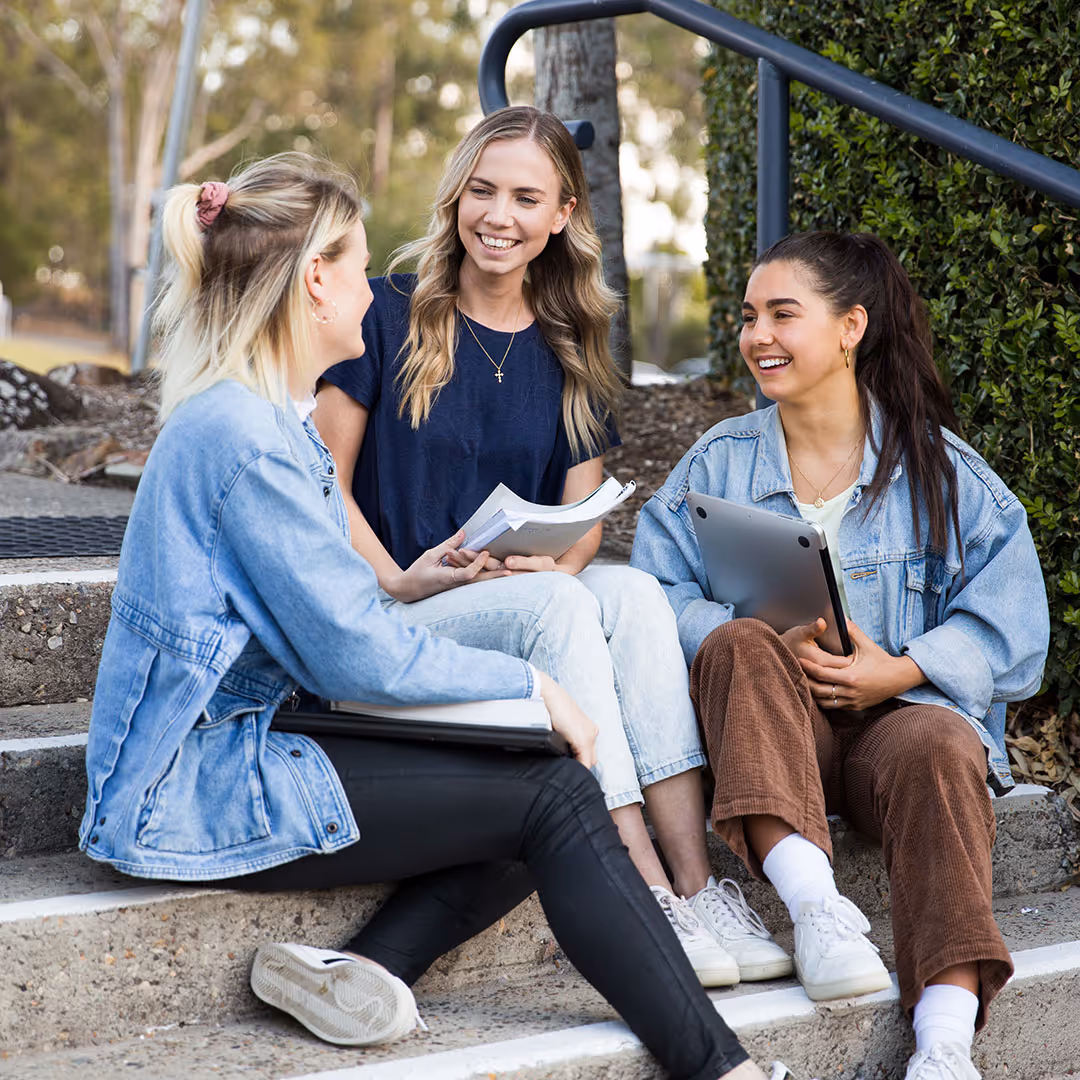 Three young women sitting on outdoor steps, smiling and holding notebooks and a laptop while chatting.