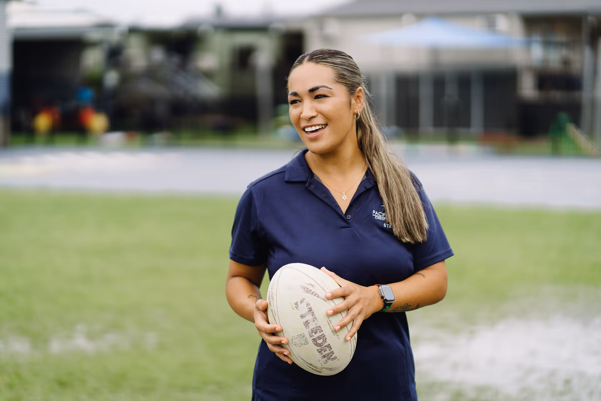 Woman smiling and holding a rugby ball on a grassy sports field.