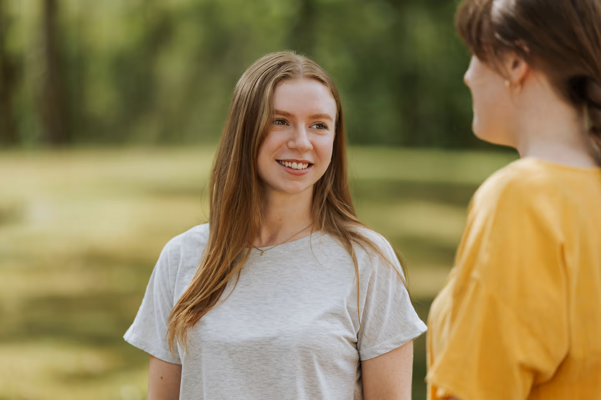 Smiling young woman with long hair wearing a light gray t-shirt, talking to another person in a yellow top outdoors.