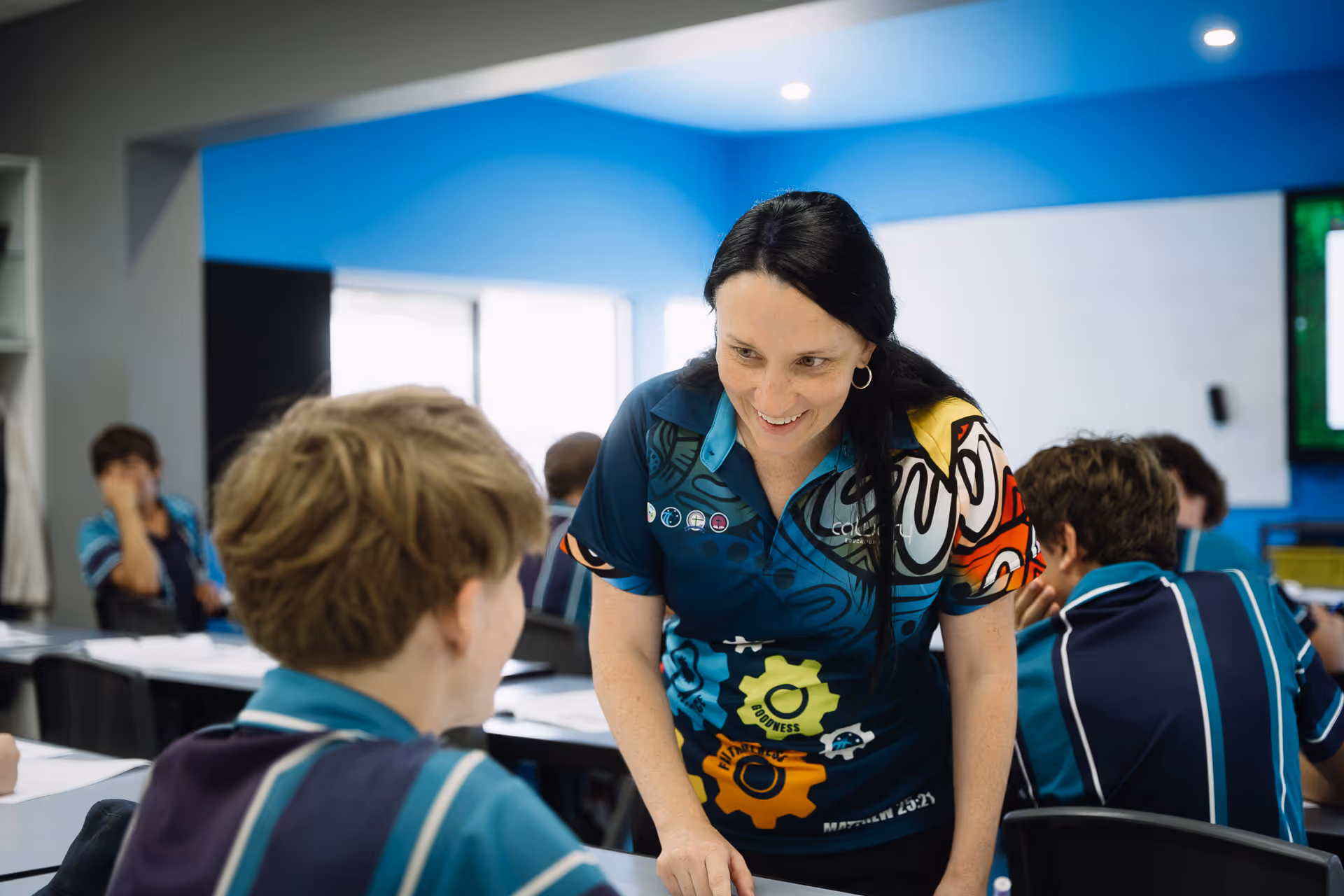 Teacher smiling and engaging with a student in a classroom with other students seated at desks.