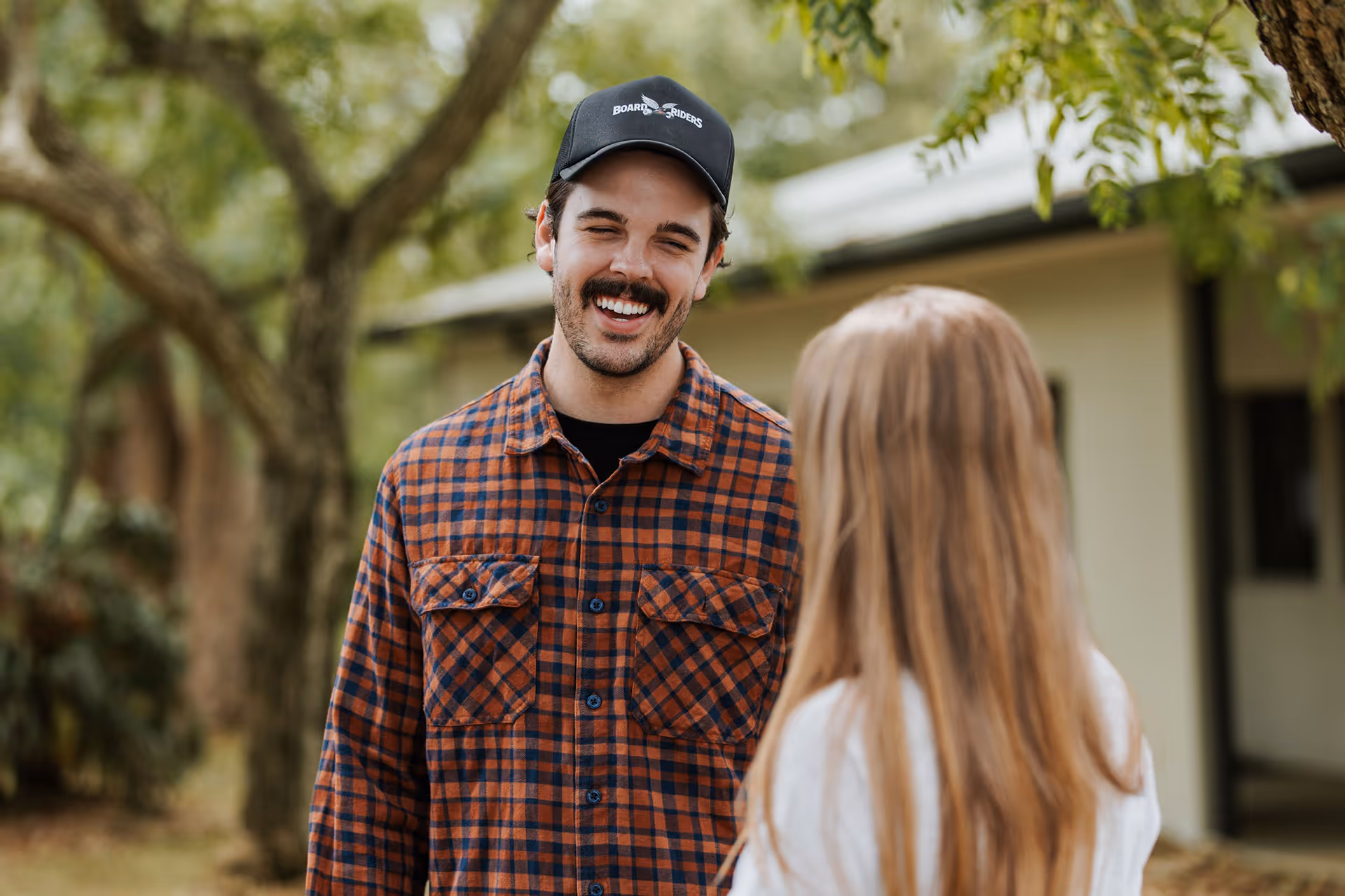 Man wearing a black cap and orange plaid shirt smiling at a woman with long blonde hair outdoors.