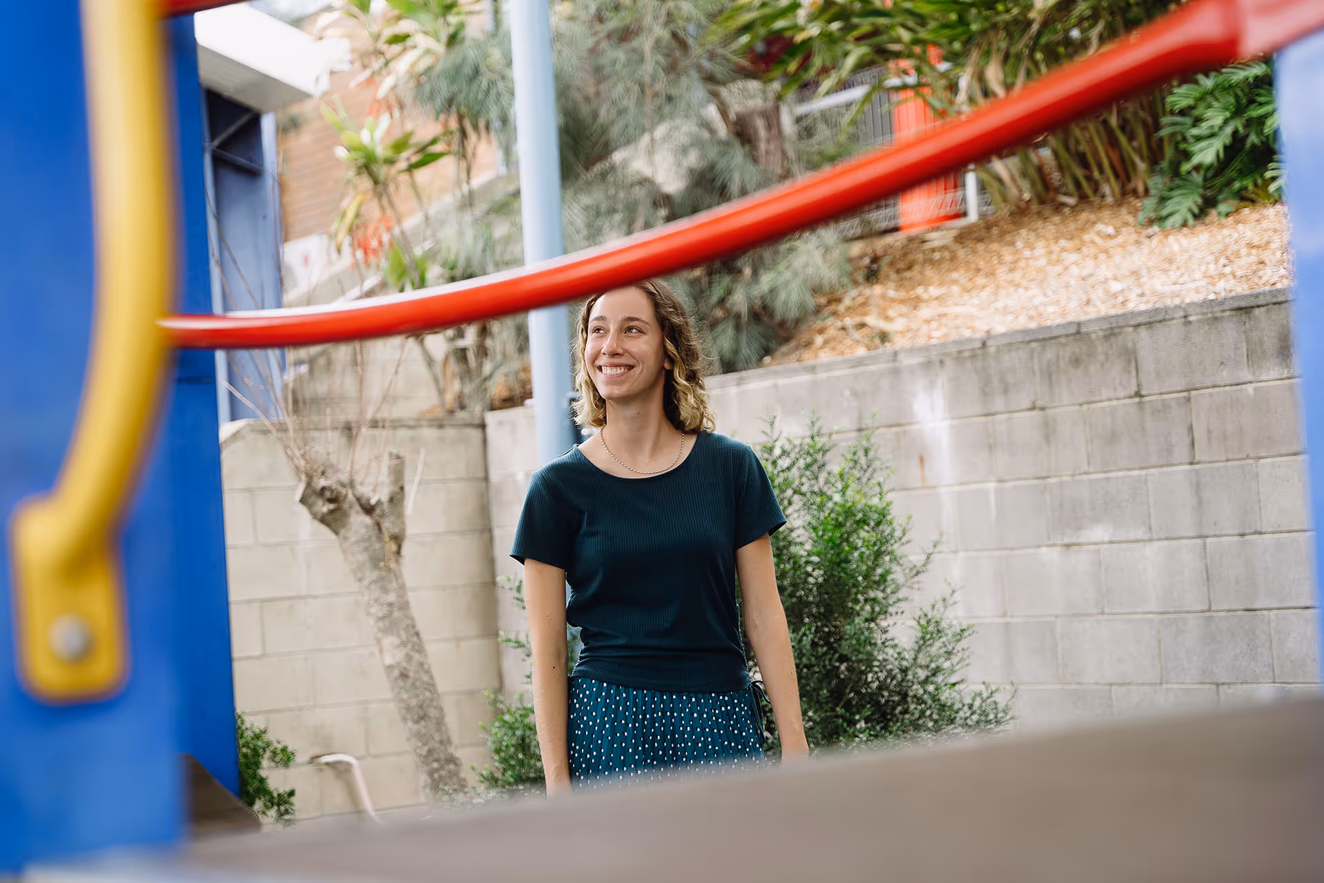 Smiling woman in a black shirt and blue skirt standing outdoors near a concrete wall with plants and playground equipment in the foreground.
