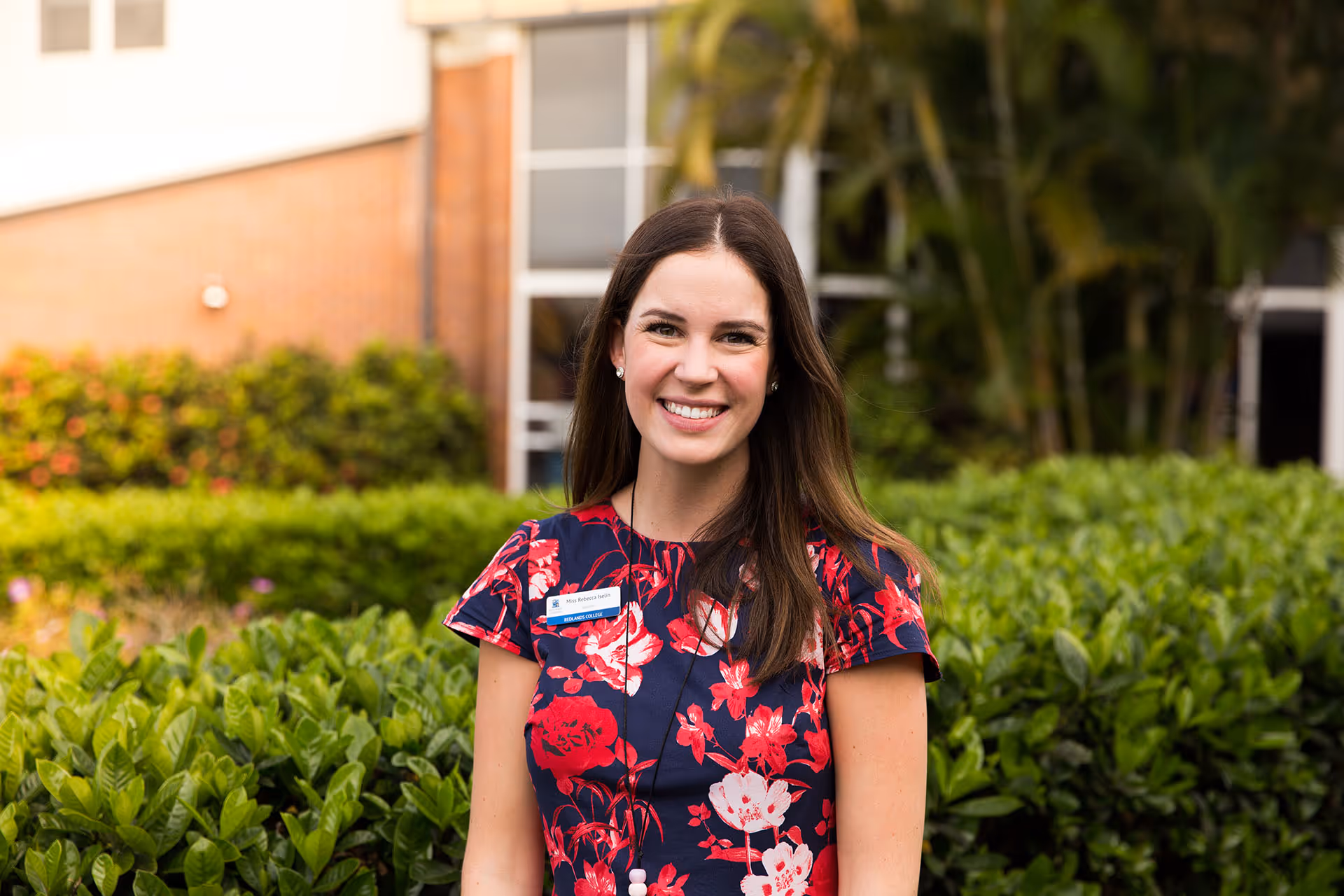 Smiling woman with long brown hair wearing a navy blue dress with red and white floral patterns standing in front of green bushes and a building.