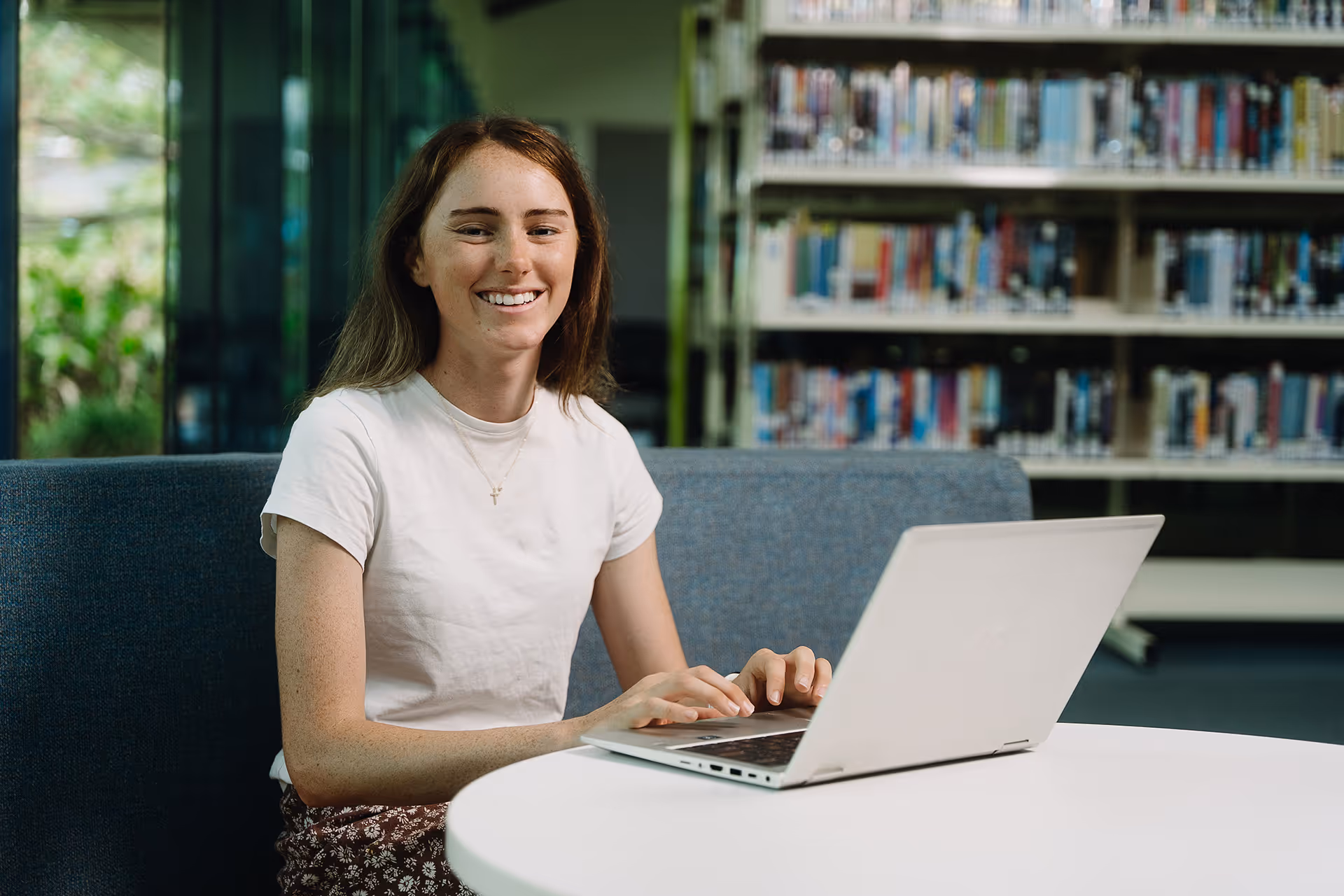 Smiling young woman typing on a laptop in a library with shelves of books in the background.