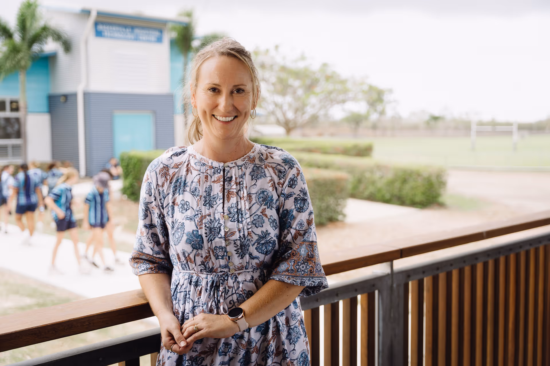 Smiling woman in a floral dress standing on a balcony with a school building and children in uniforms in the background.