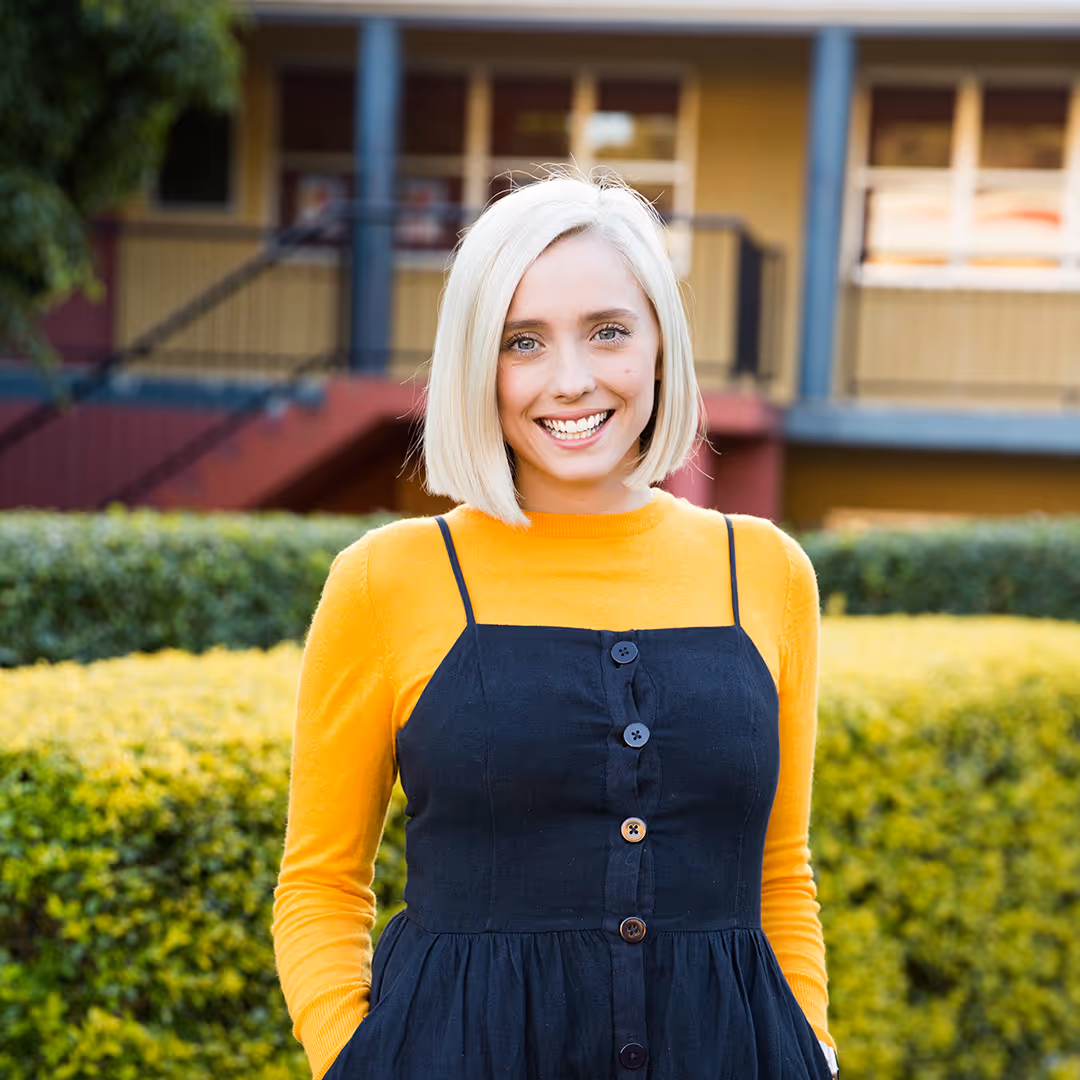 Smiling blonde woman wearing a black dress over a yellow long-sleeve top standing outdoors with green bushes and a building in the background.