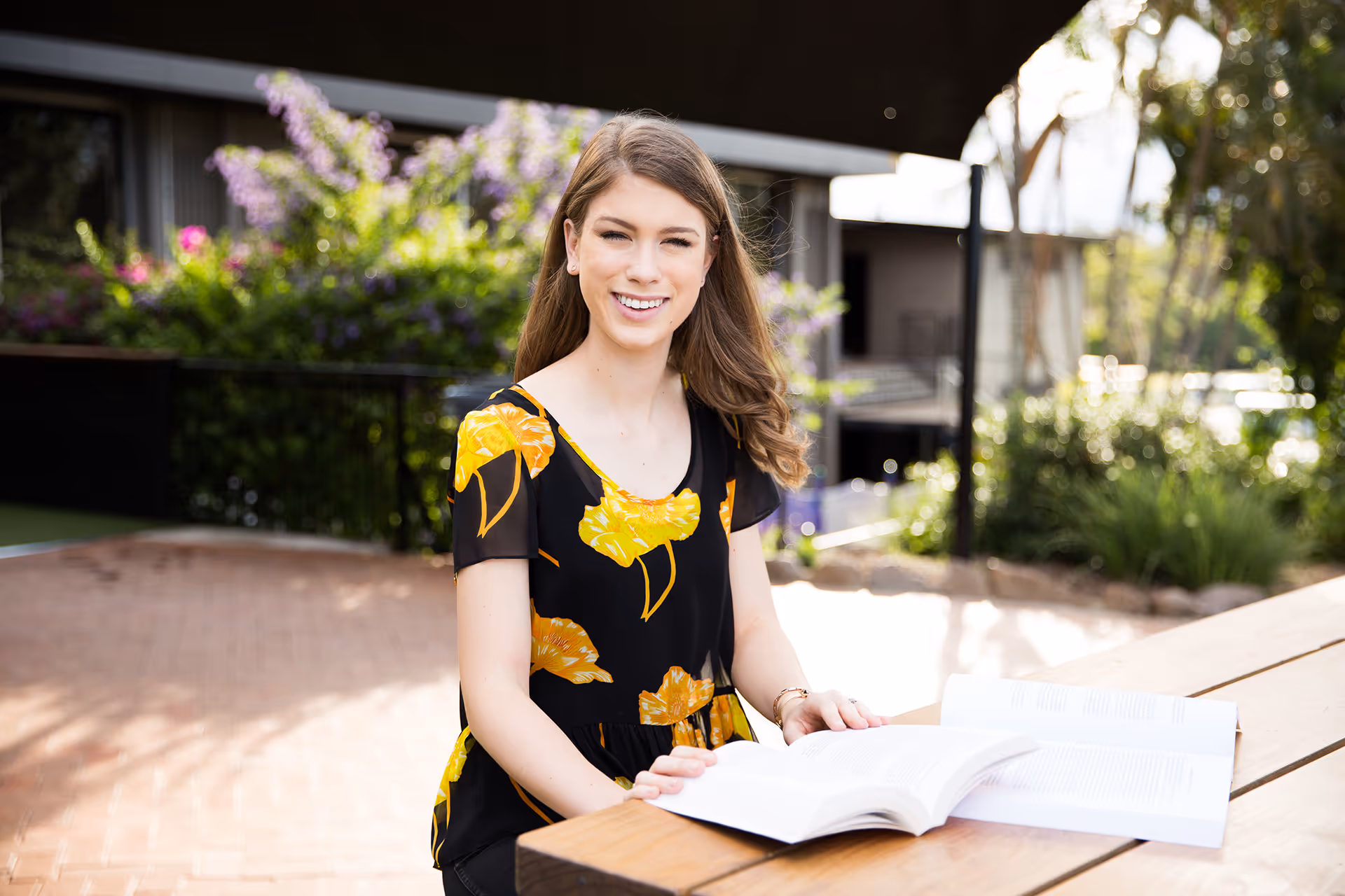 Smiling young woman in floral black and yellow top sitting at an outdoor wooden table reading a book.