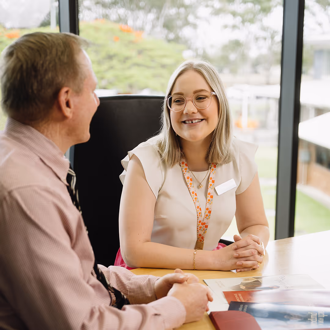 Smiling woman with glasses and name tag talking to a man at a table with brochures in a bright office.