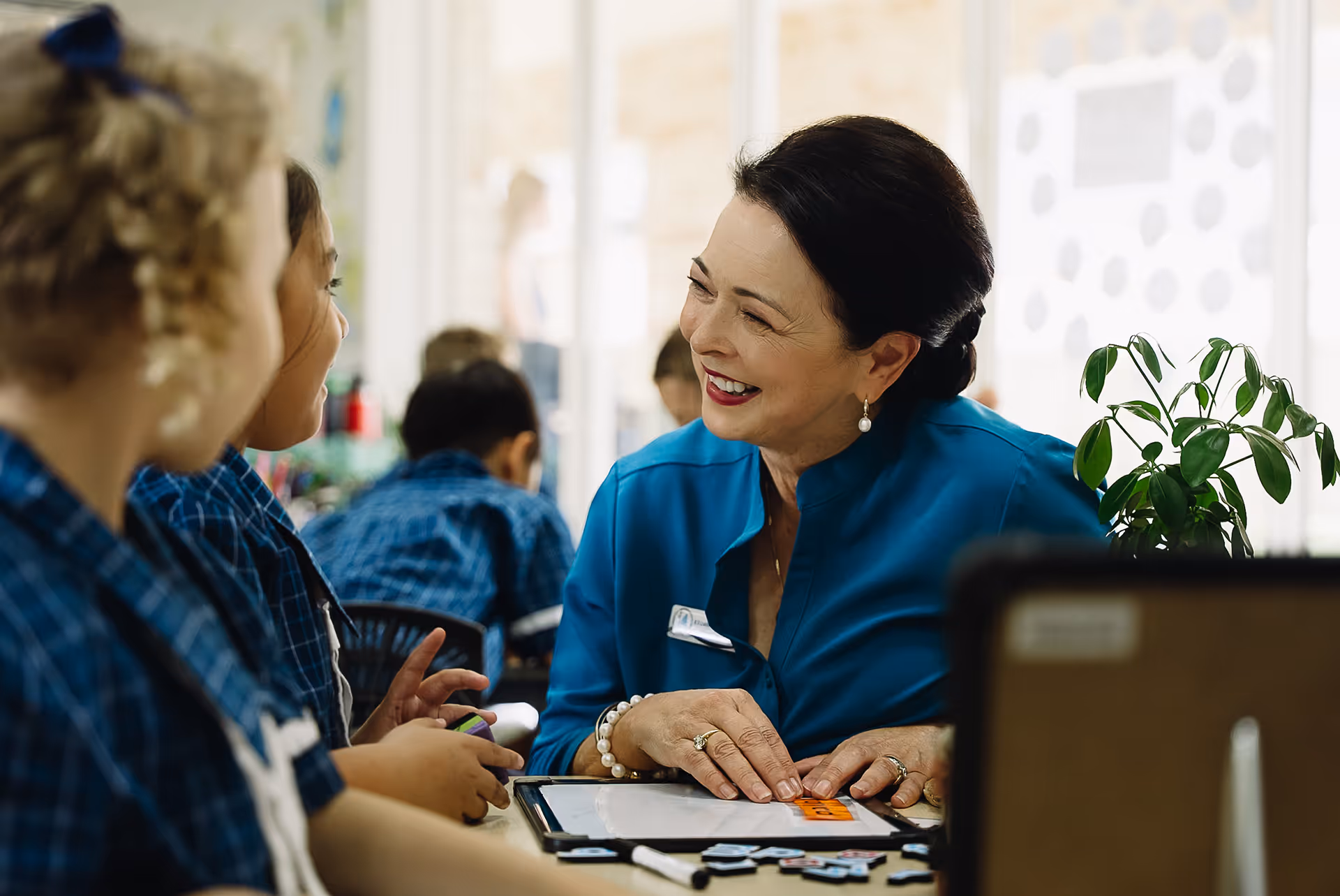 Teacher in blue blouse smiling and interacting with young students at a classroom table with educational materials.