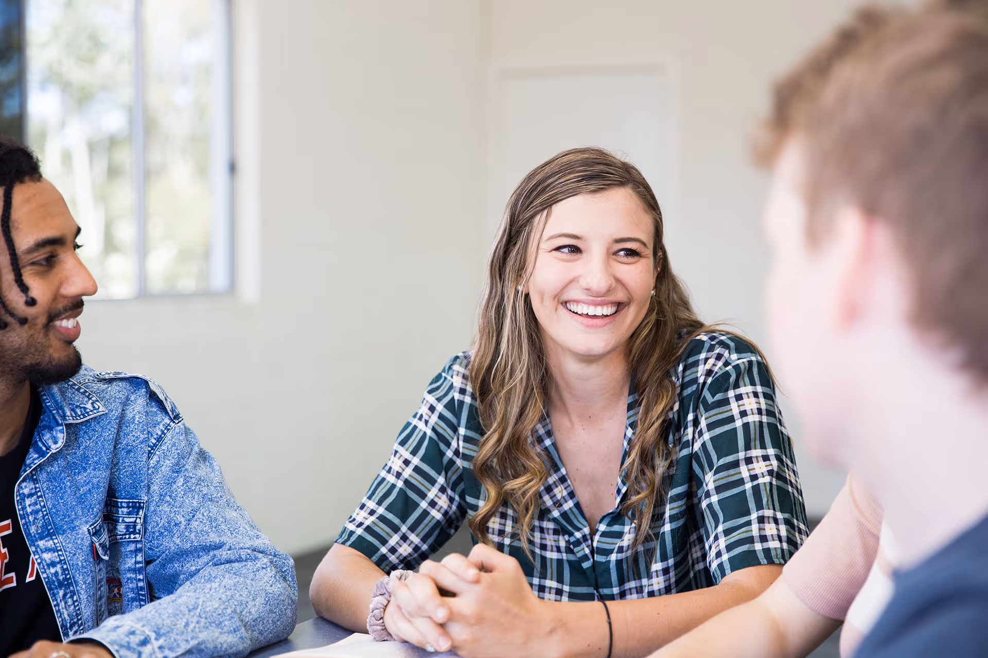Three young adults sitting indoors, smiling and engaged in conversation, with a woman in a green plaid shirt at the center.