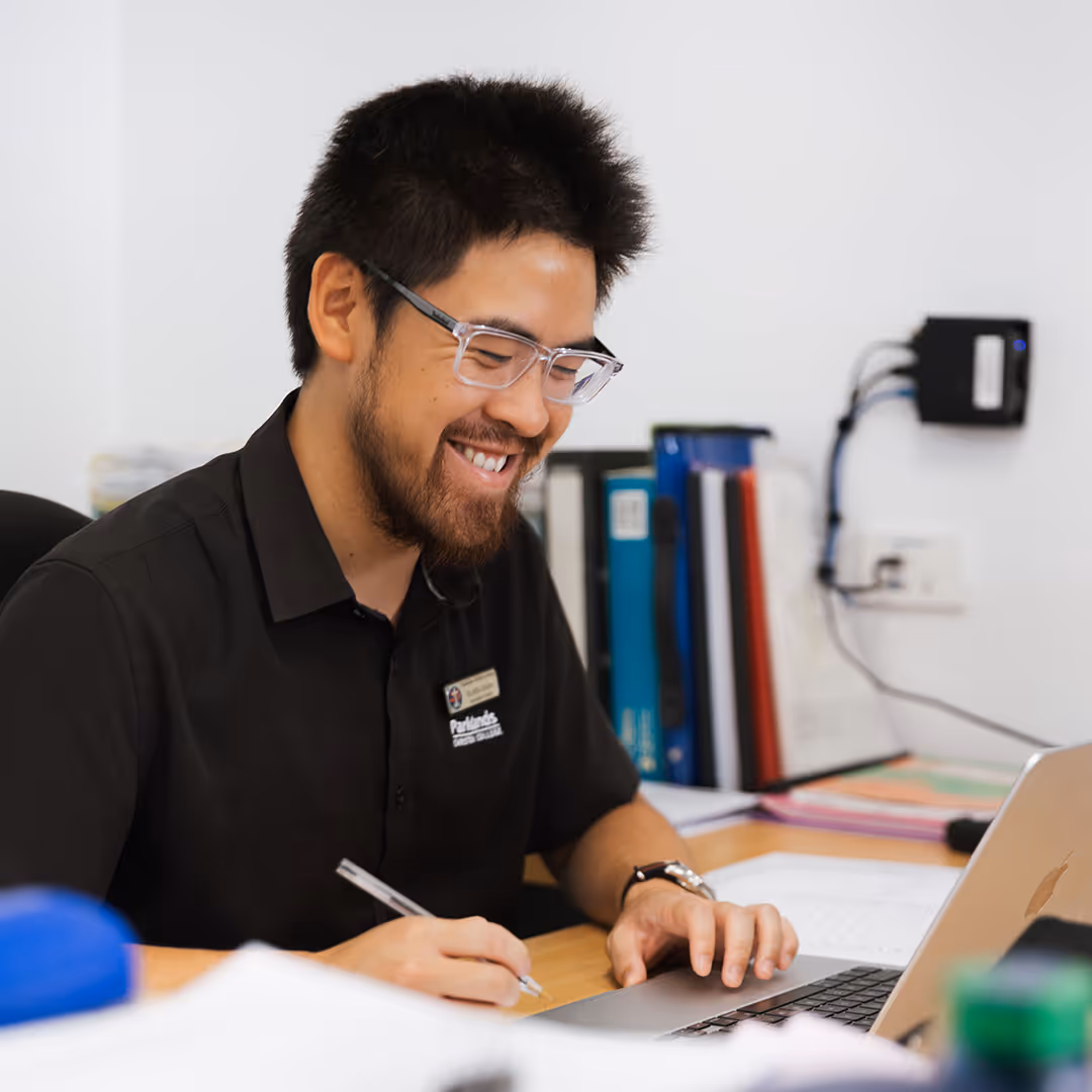 Smiling man with glasses writing with a pen and using a laptop at a desk with folders in the background.