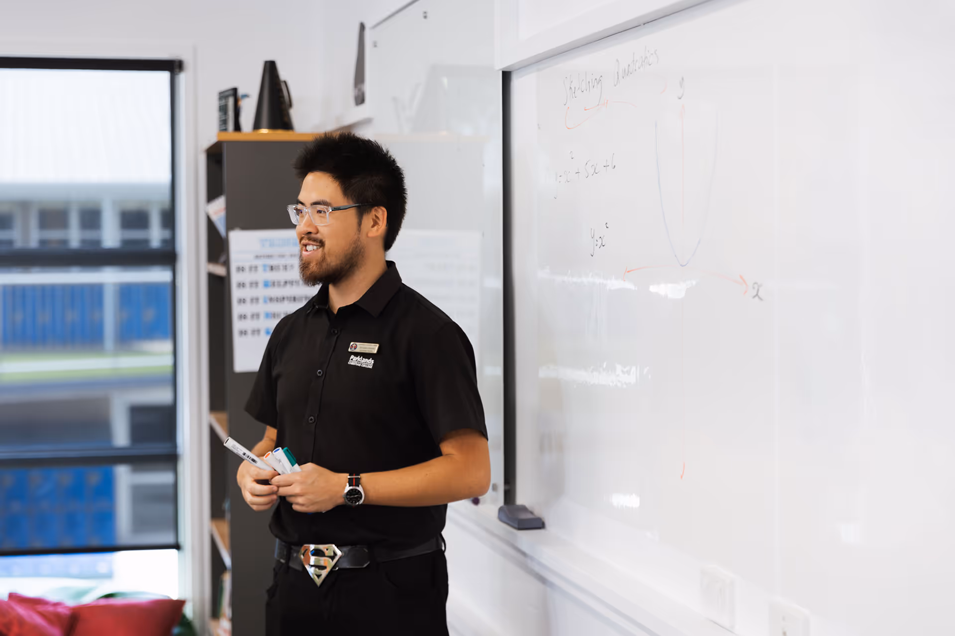 Teacher holding markers and standing next to a whiteboard with math equations and a graph.