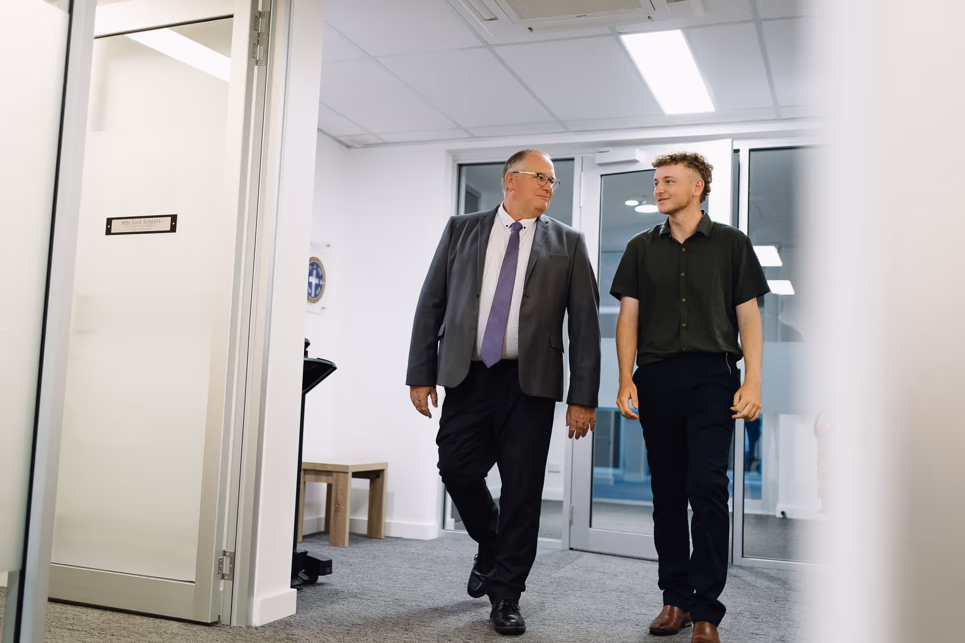 Two men walking and talking in a modern office hallway, one in a suit and tie, the other in casual shirt and pants.
