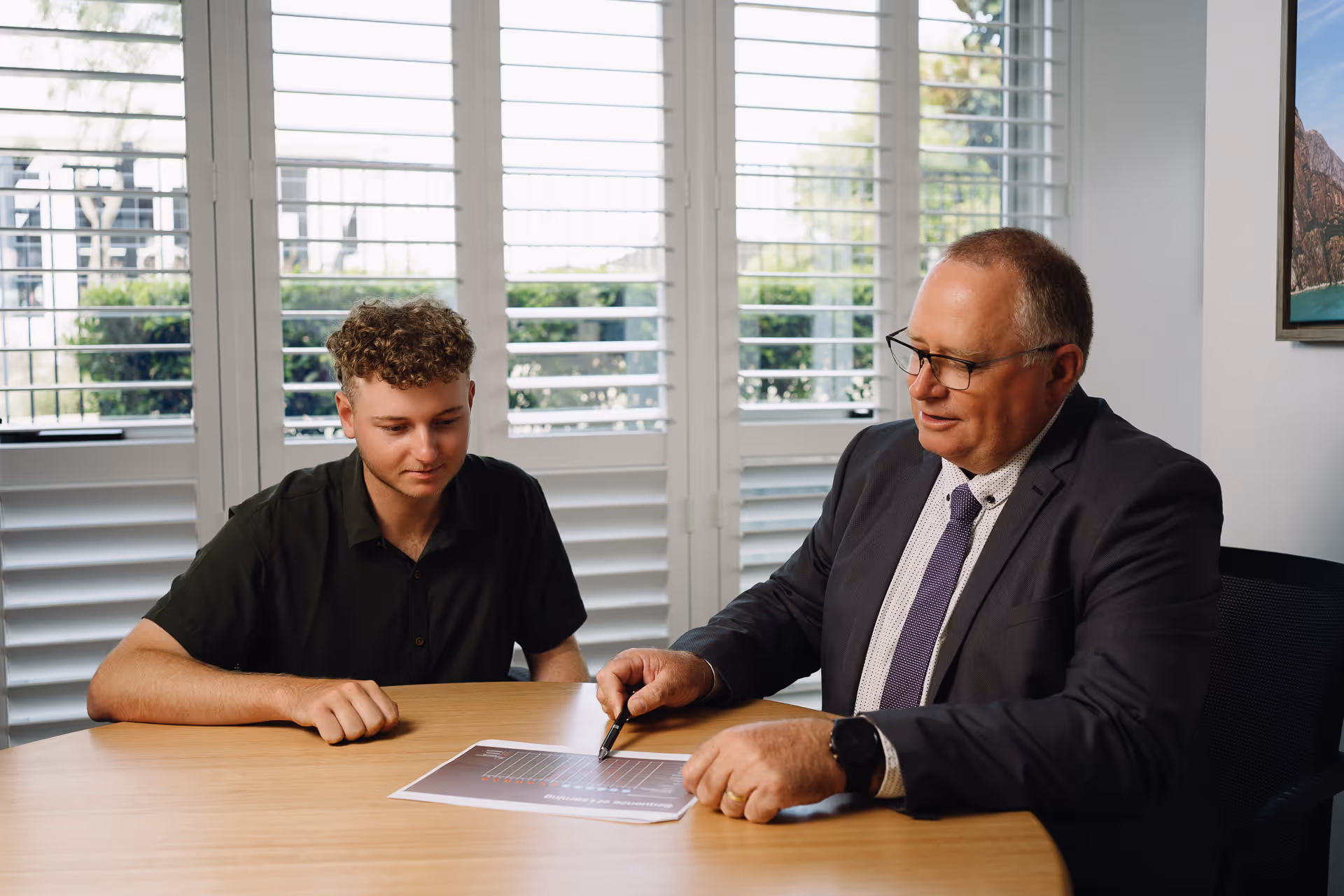 A professional man in a suit explains a chart to a young man sitting across the table in a bright office.