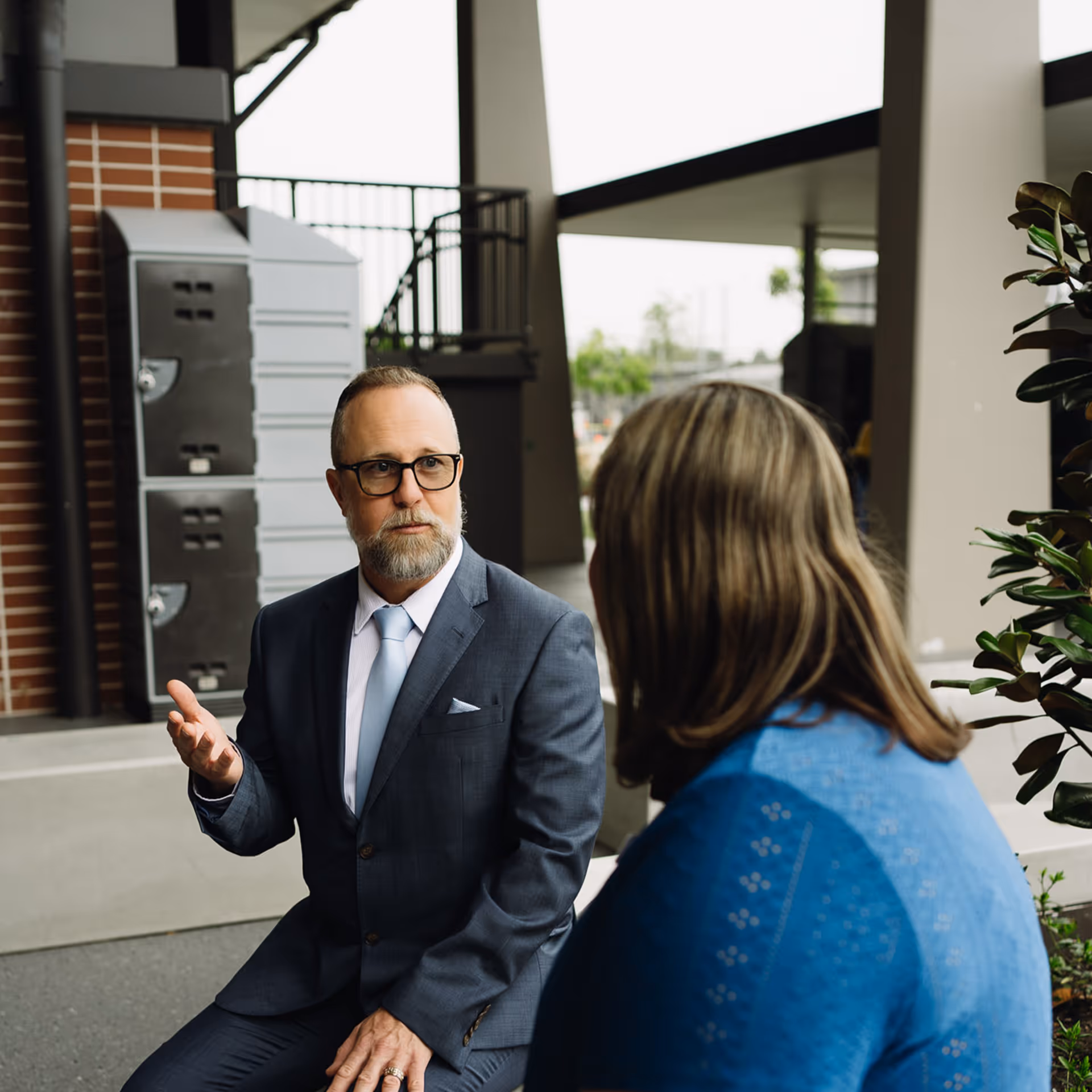 Man in a gray suit and glasses talking to a woman in a blue sweater outdoors.