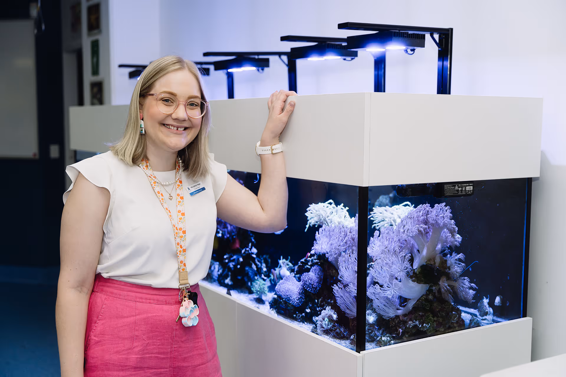 Smiling woman with glasses and pink skirt standing beside a large illuminated aquarium with coral inside.