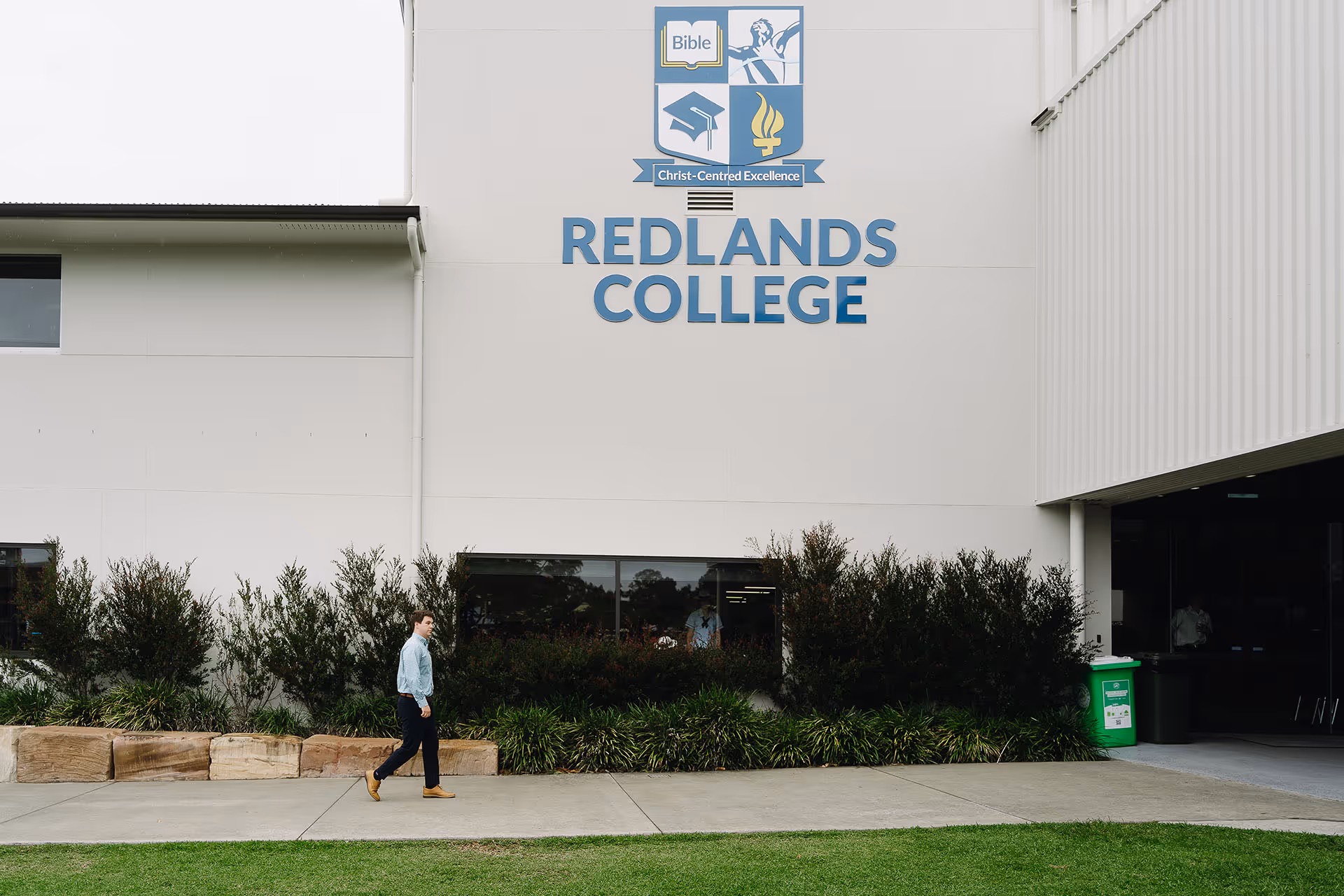 Person walking on sidewalk in front of Redlands College building with logo and greenery.