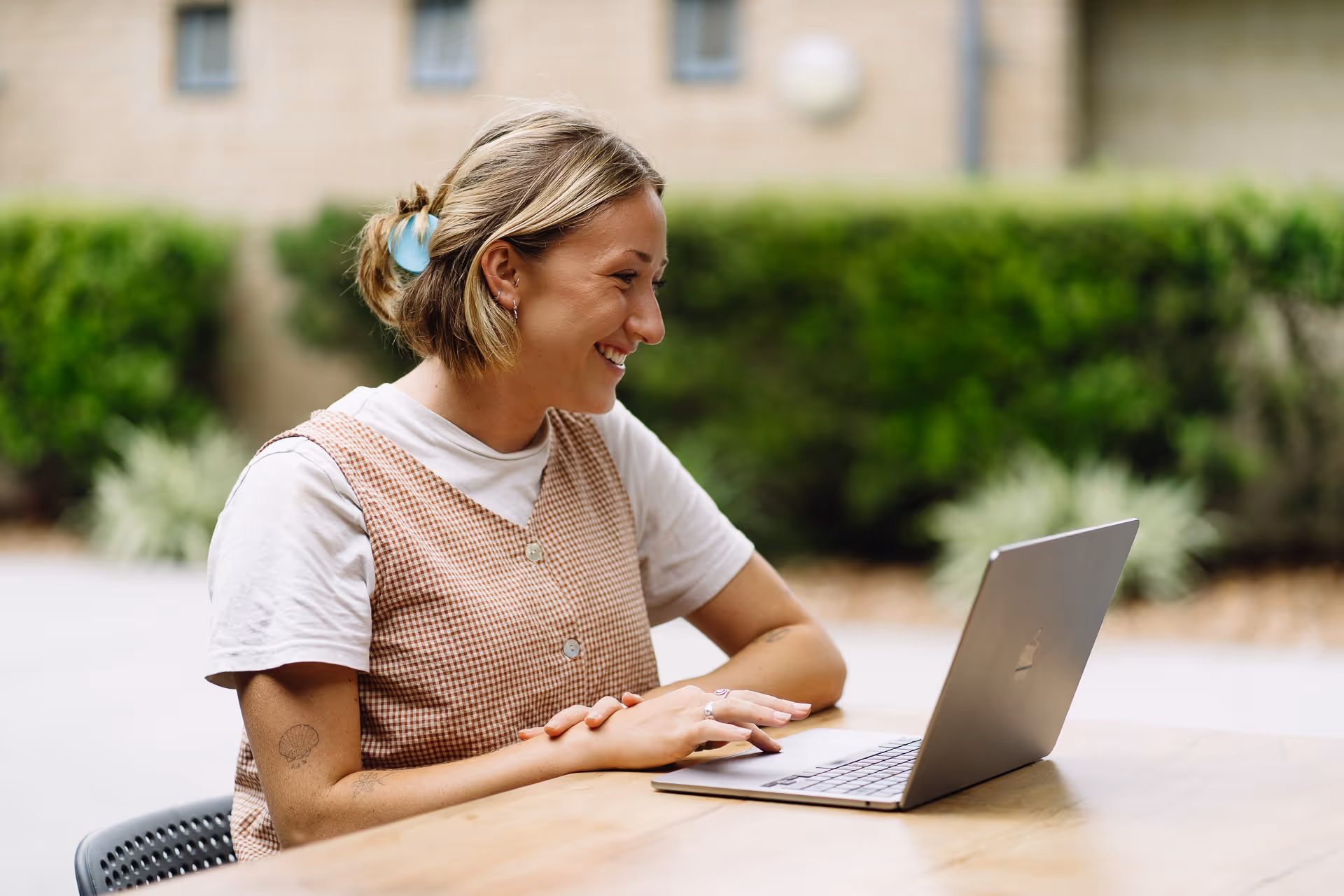 Smiling woman with short blond hair, wearing a checkered vest and white shirt, using a laptop outdoors at a wooden table.