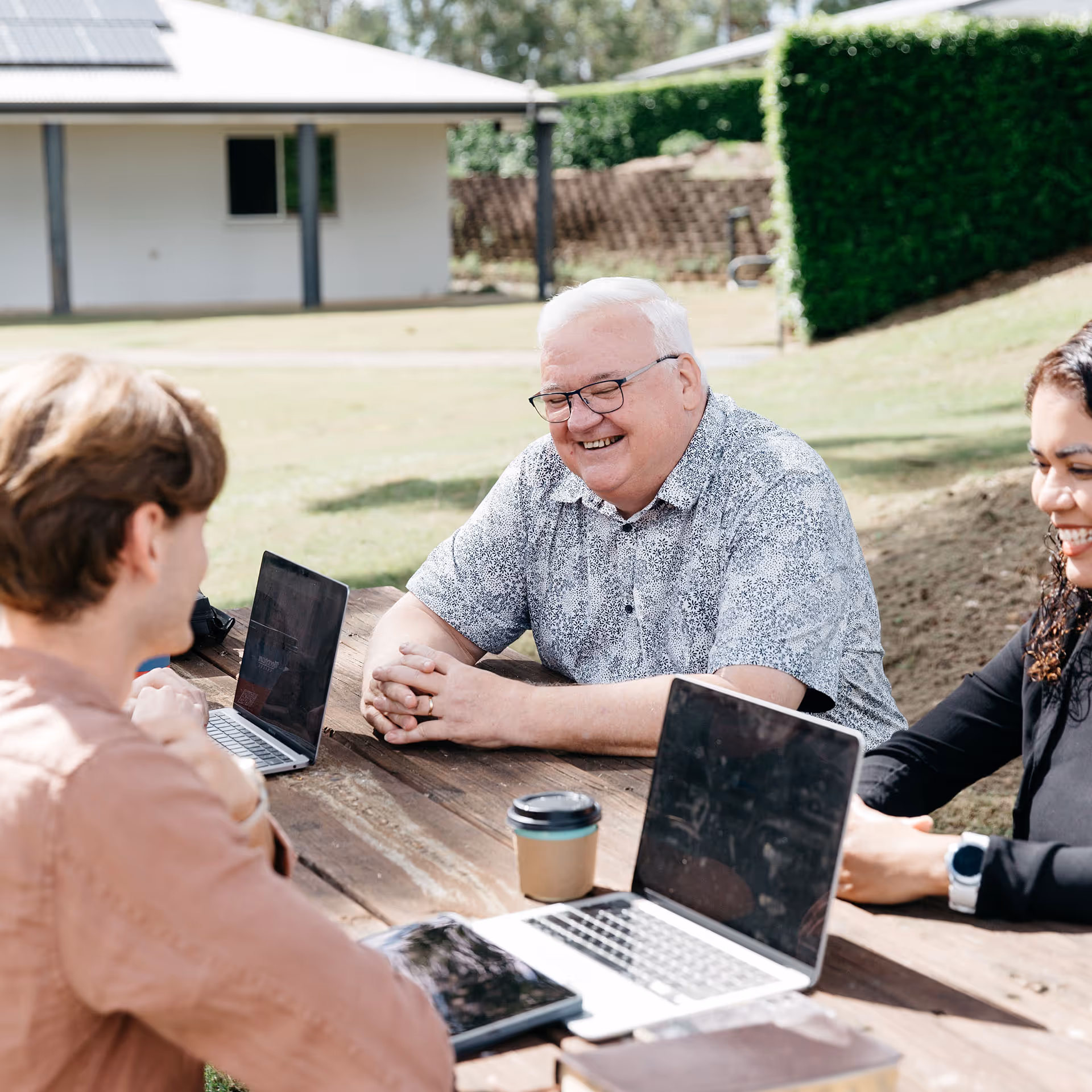 Three people sitting outdoors at a wooden table with laptops, smiling and engaging in conversation.
