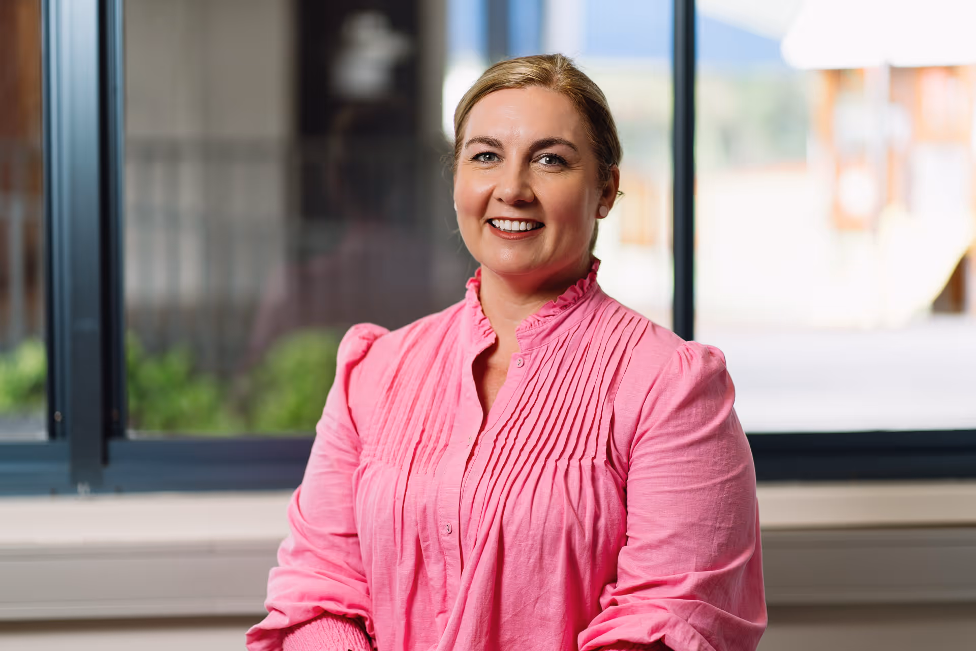 Smiling woman with blonde hair wearing a pink blouse, sitting indoors near a window.