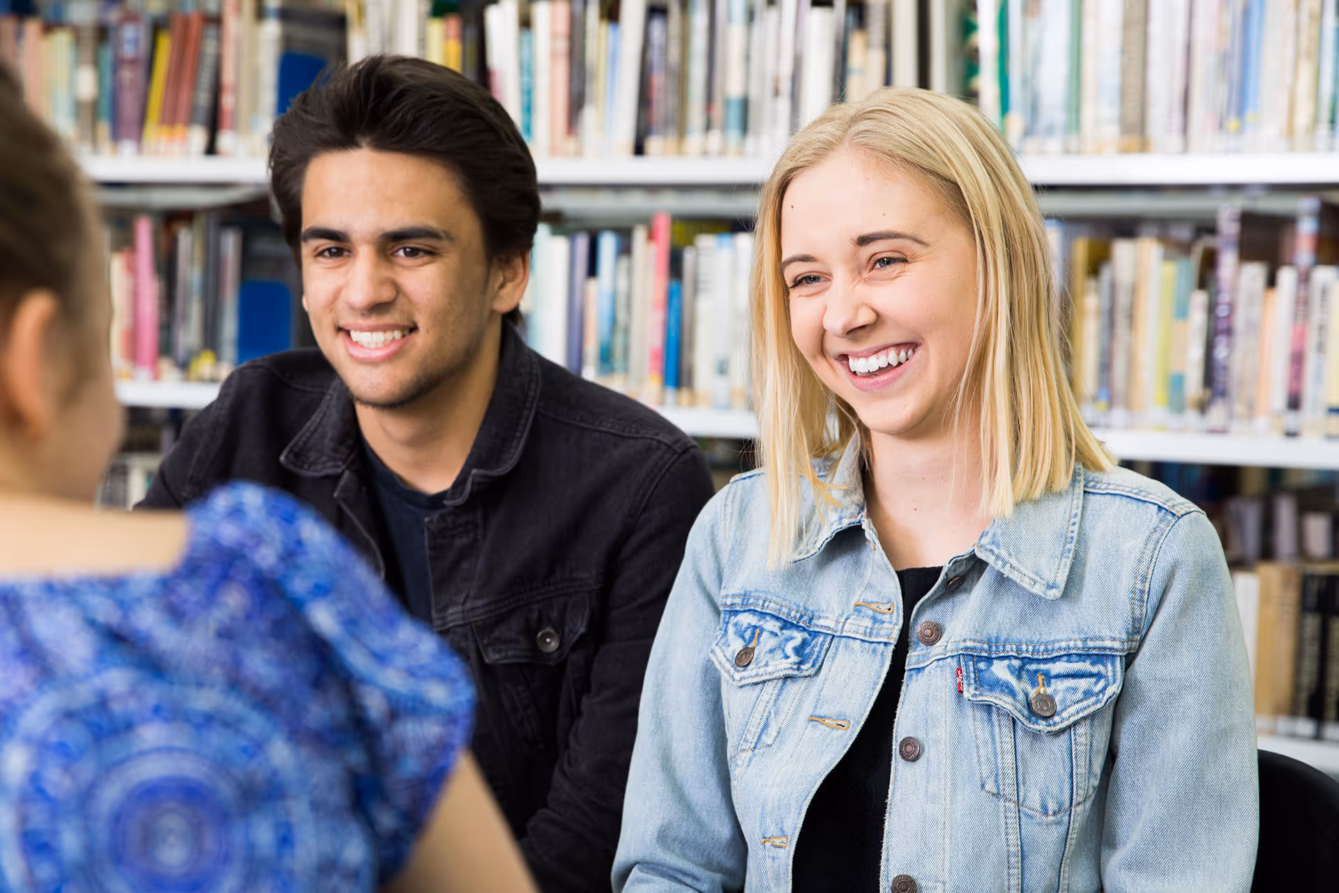 Two young adults smiling and talking in a library with bookshelves in the background.