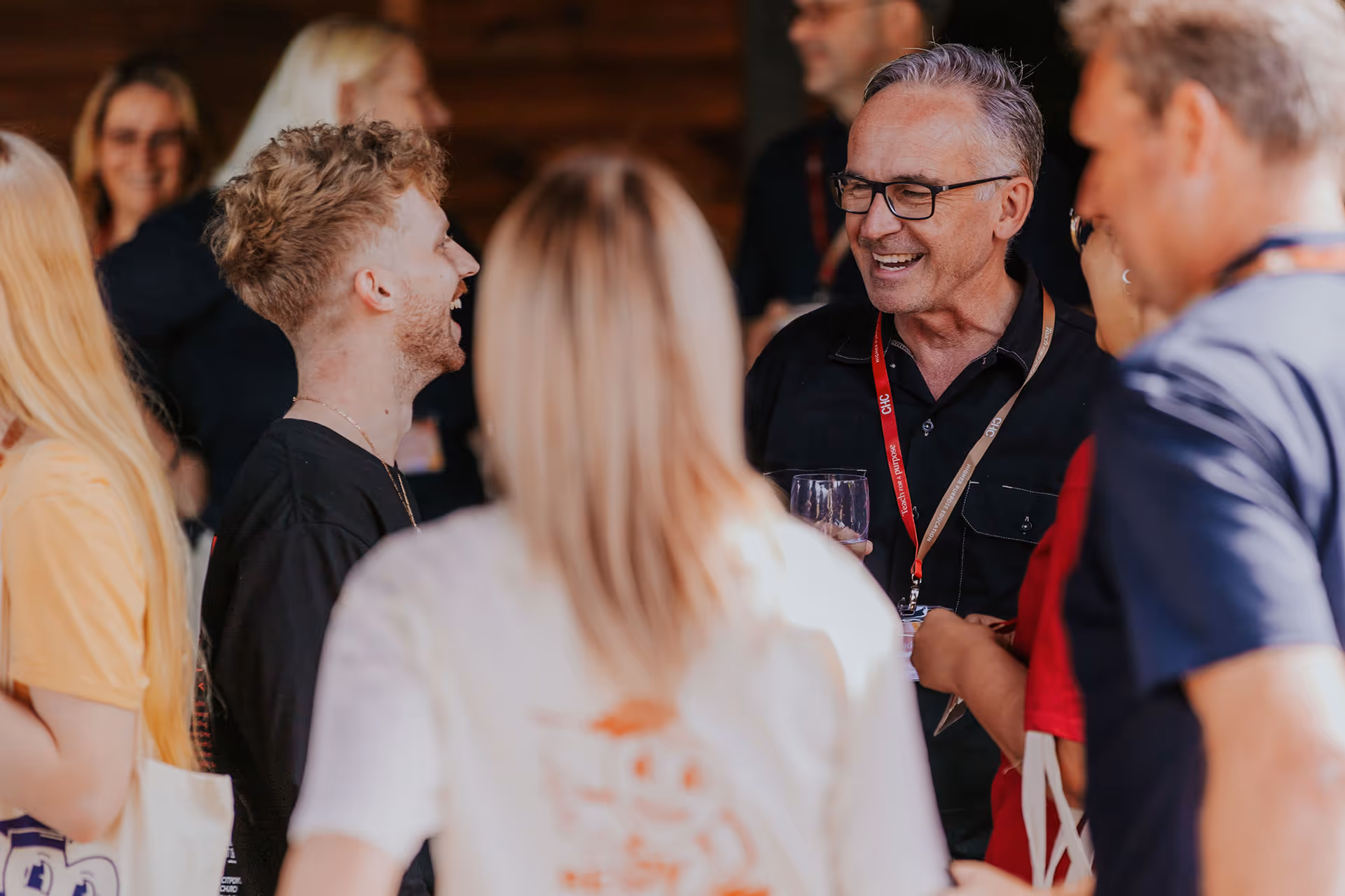 Group of people smiling and socializing indoors, with one man holding a glass and wearing a red lanyard.