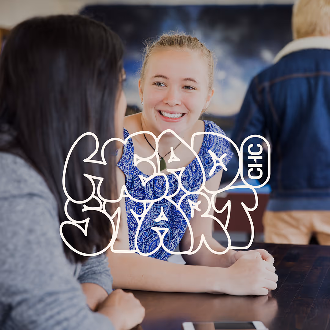 Two women sitting at a table, smiling and conversing, with a person standing in the background.