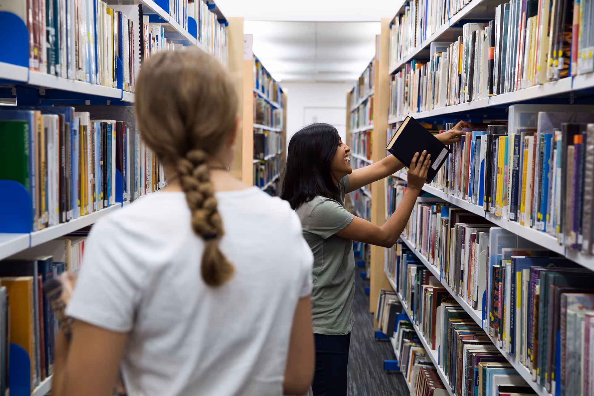 Two women browsing books in a well-lit library aisle filled with shelves of books.