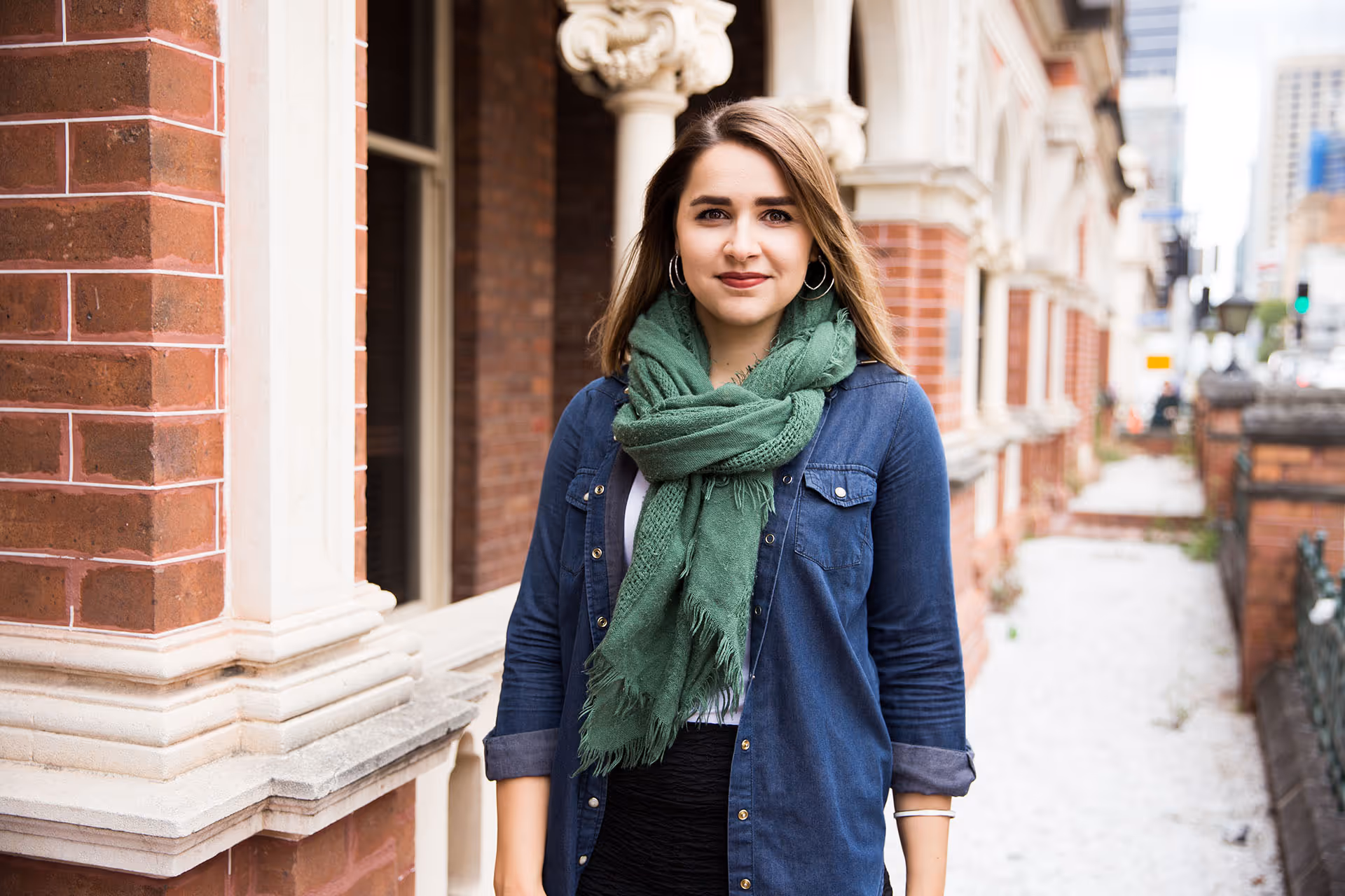 Young woman with long brown hair wearing a green scarf and denim jacket standing near a brick building in an urban setting.