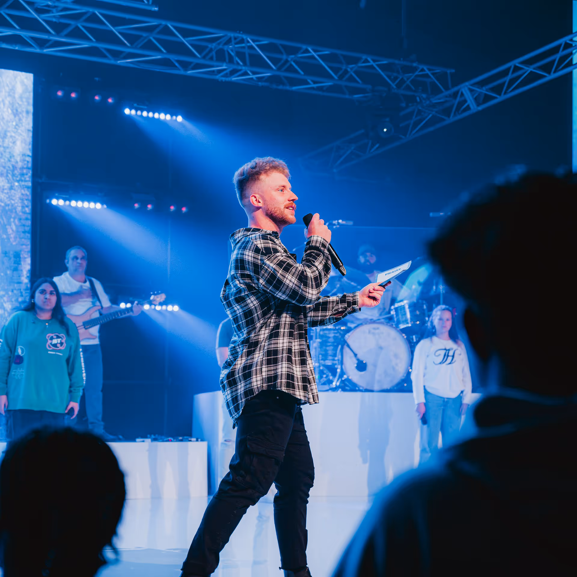Man in checkered shirt speaking into a microphone on stage with musicians and singers in the background under blue lighting.