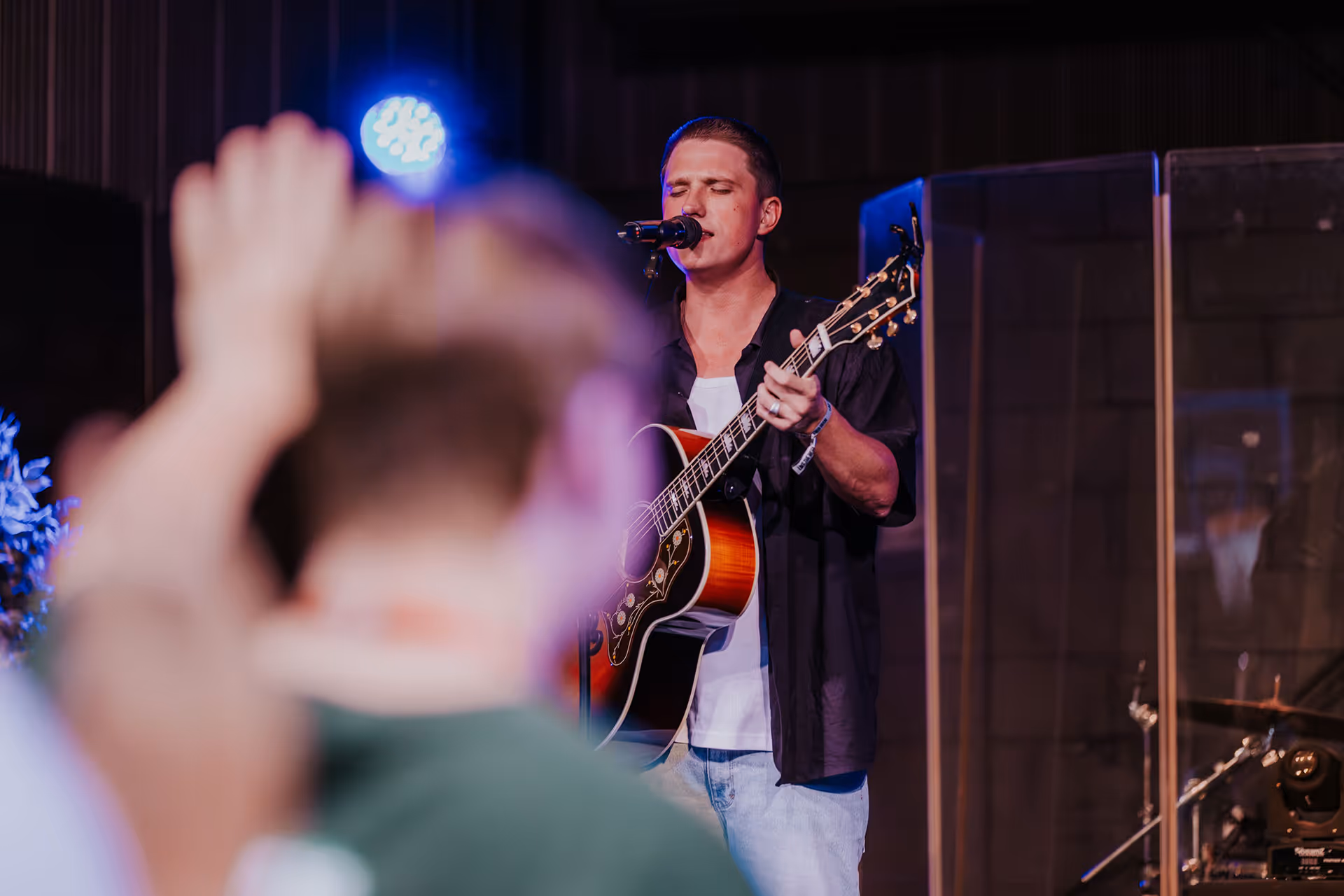 Man singing and playing acoustic guitar on stage with an audience member raising a hand in the foreground.