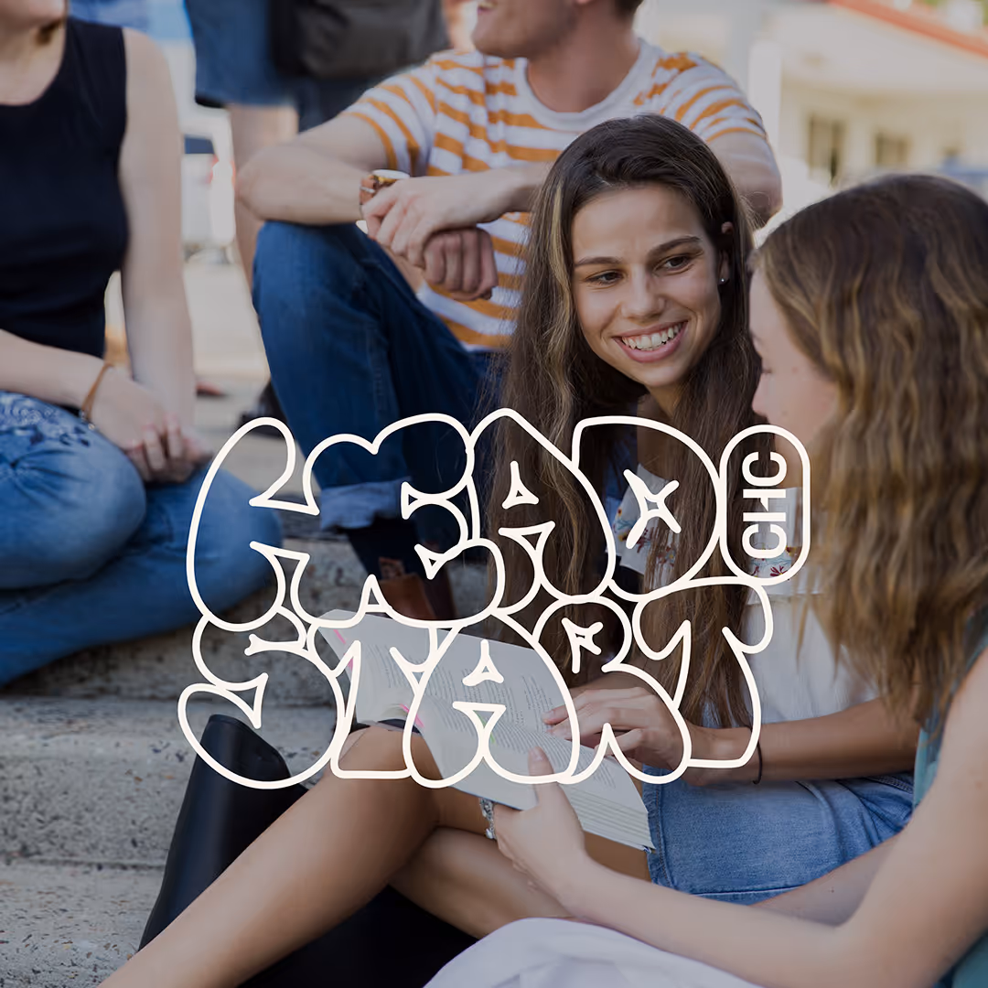 Two young women sitting on outdoor steps, smiling and reading a book together, with other people in the background.