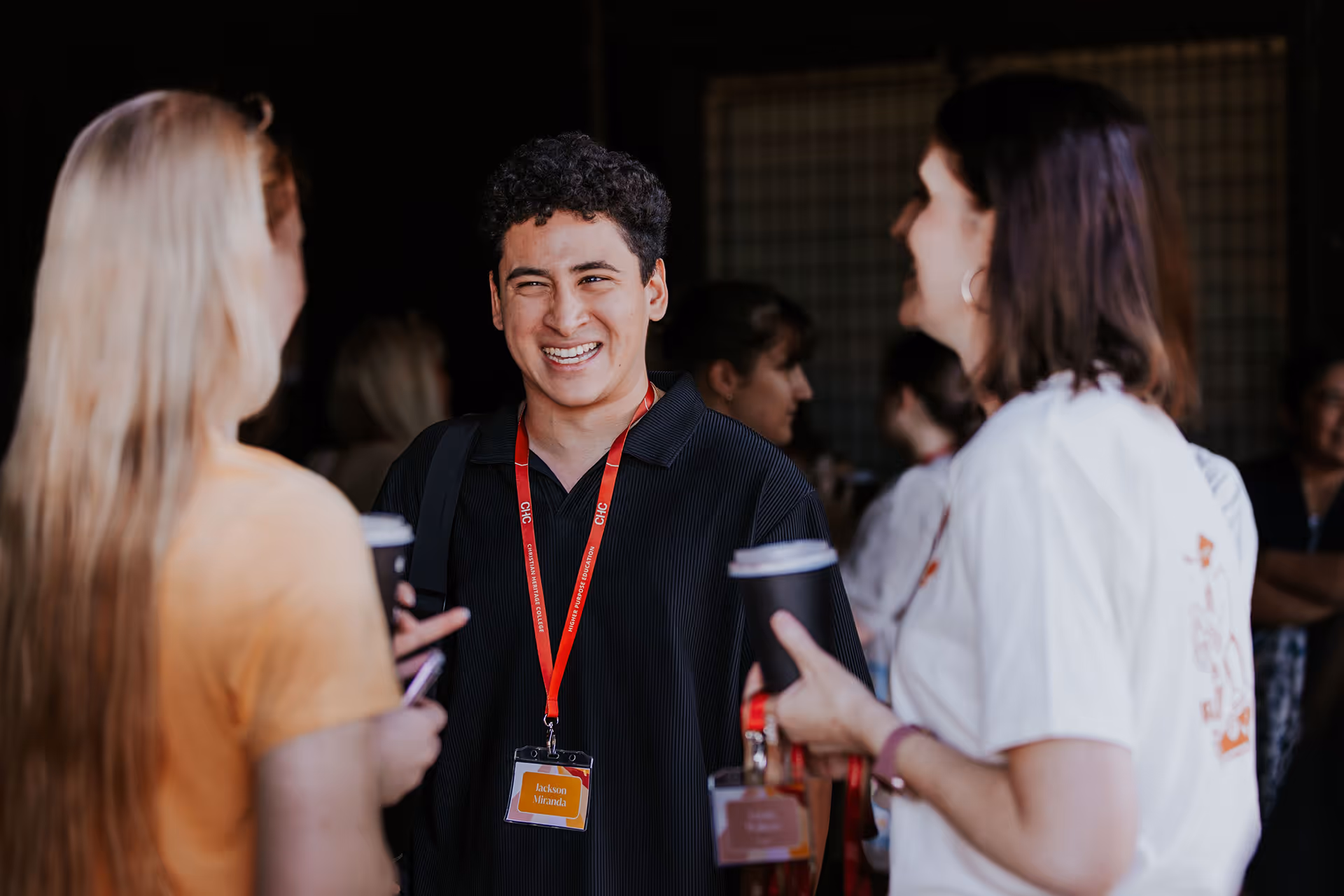 A young man with a red lanyard and name tag smiling and talking with two women holding coffee cups in a social setting.