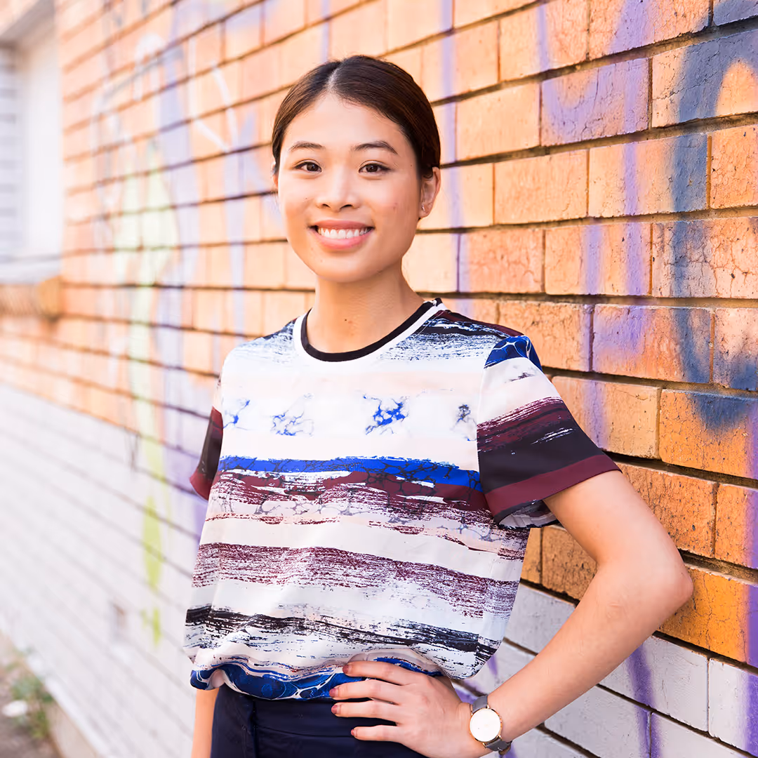 Smiling young woman with neatly tied hair wearing a striped shirt standing confidently against a graffiti-covered brick wall.